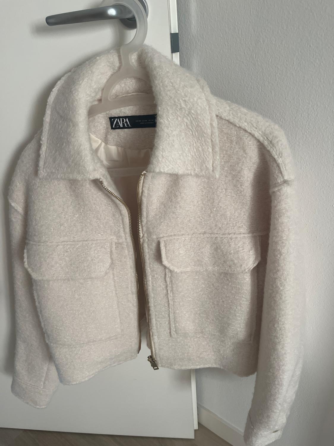Ecru textured jacket with a zip and pockets, hanging from a door handle. Text visible on the label: Zara.