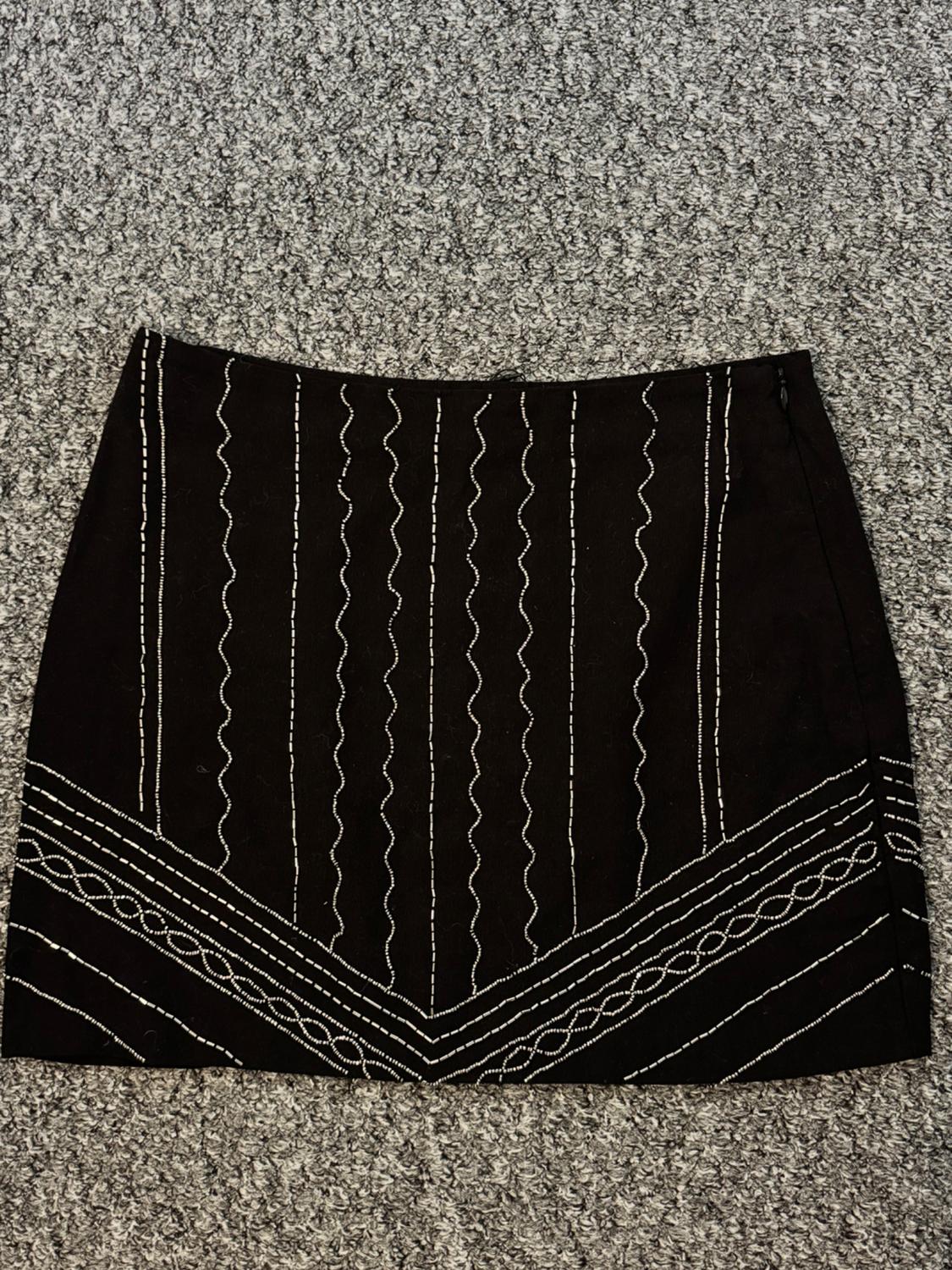 Short black skirt with white embroidery in geometric patterns and wavy lines, on a textured grey carpet background.