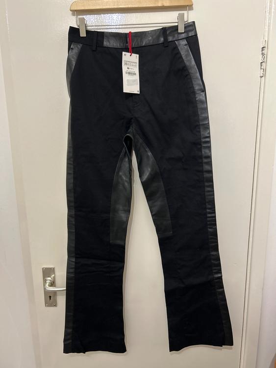 Black trousers with leather detailing on the sides, hanging on a wooden hanger. White label visible on the front.