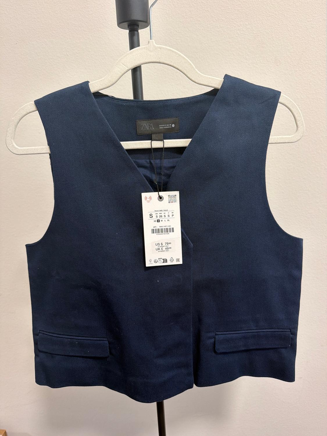 Dark blue vest hanging on a hanger with a visible price tag. White background and black metal stand.