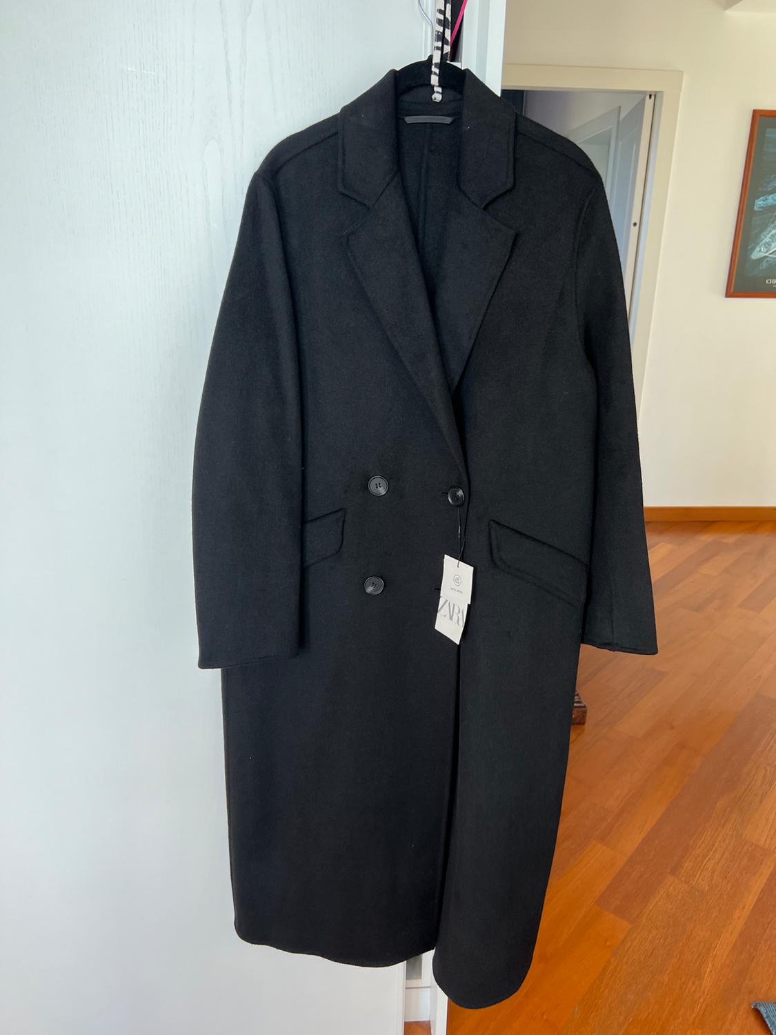 Long black double-breasted coat hanging on a white door. Interior background with wooden flooring. Visible label with the text ZARA.