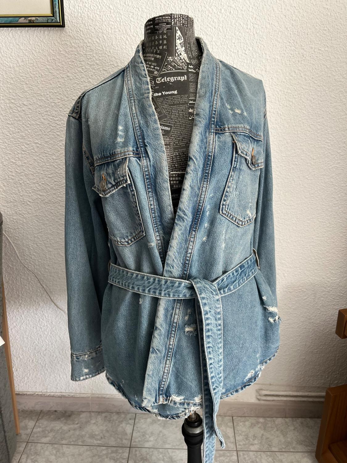 Distressed belted denim jacket on a mannequin with the text AMAZING SECRETS, Telegraph and the Young. White wall background.