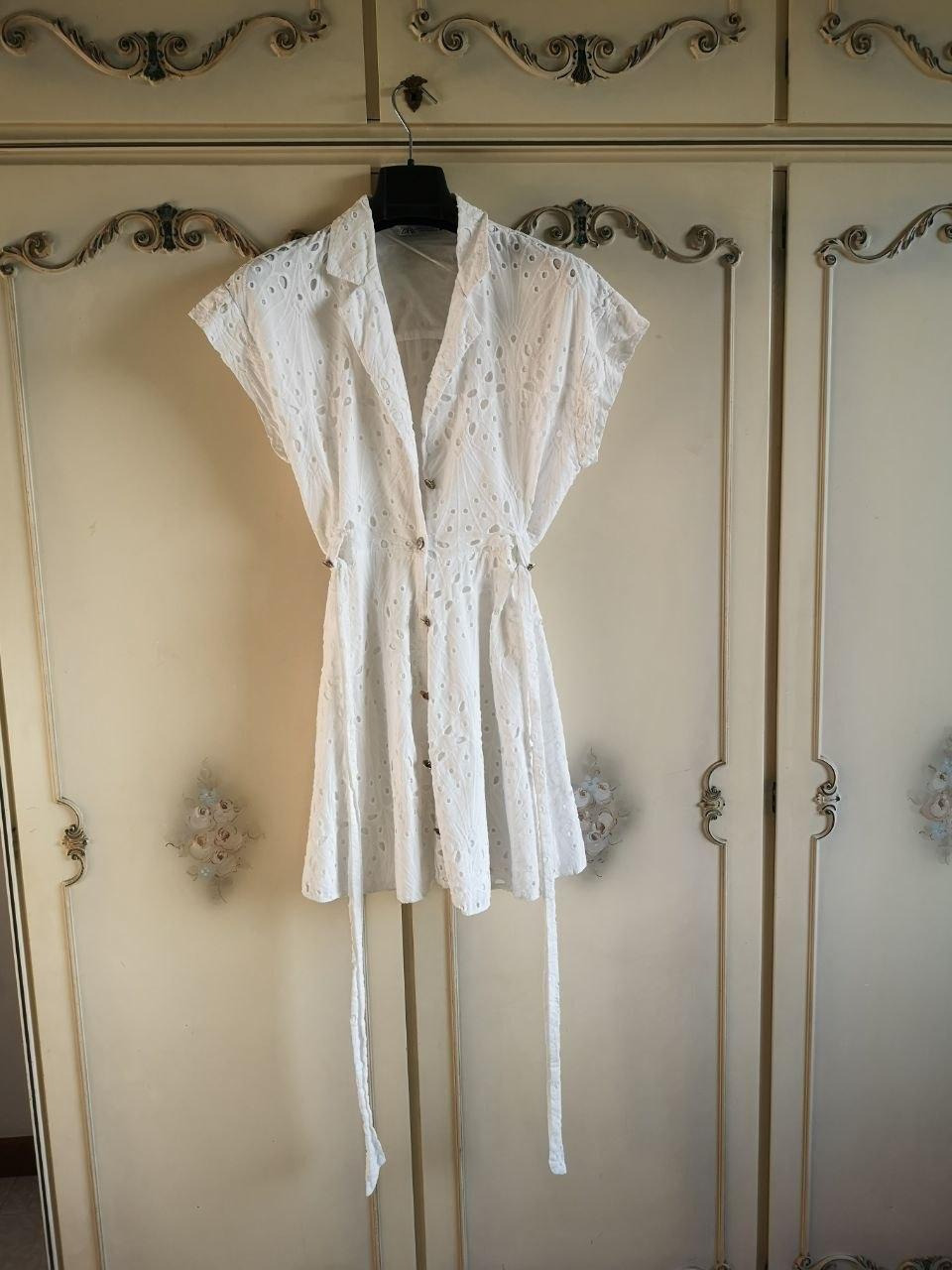 Short white broderie anglaise dress with buttons and a tie at the waist, hanging in front of a classic cream wooden wardrobe.