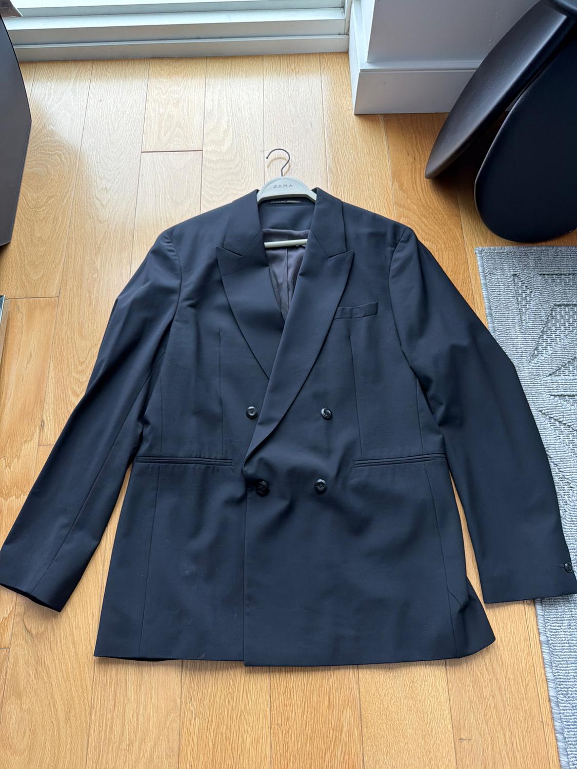 Black double-breasted suit jacket on a Zara hanger, placed on the wooden floor next to a gray rug.