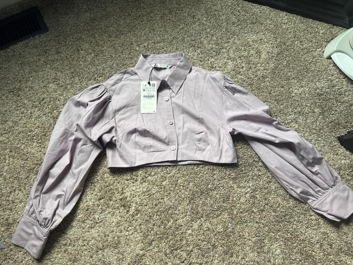 Lilac cropped shirt with a collar, buttons, and puff sleeves. The garment with a tag is laid out on a textured beige rug.
