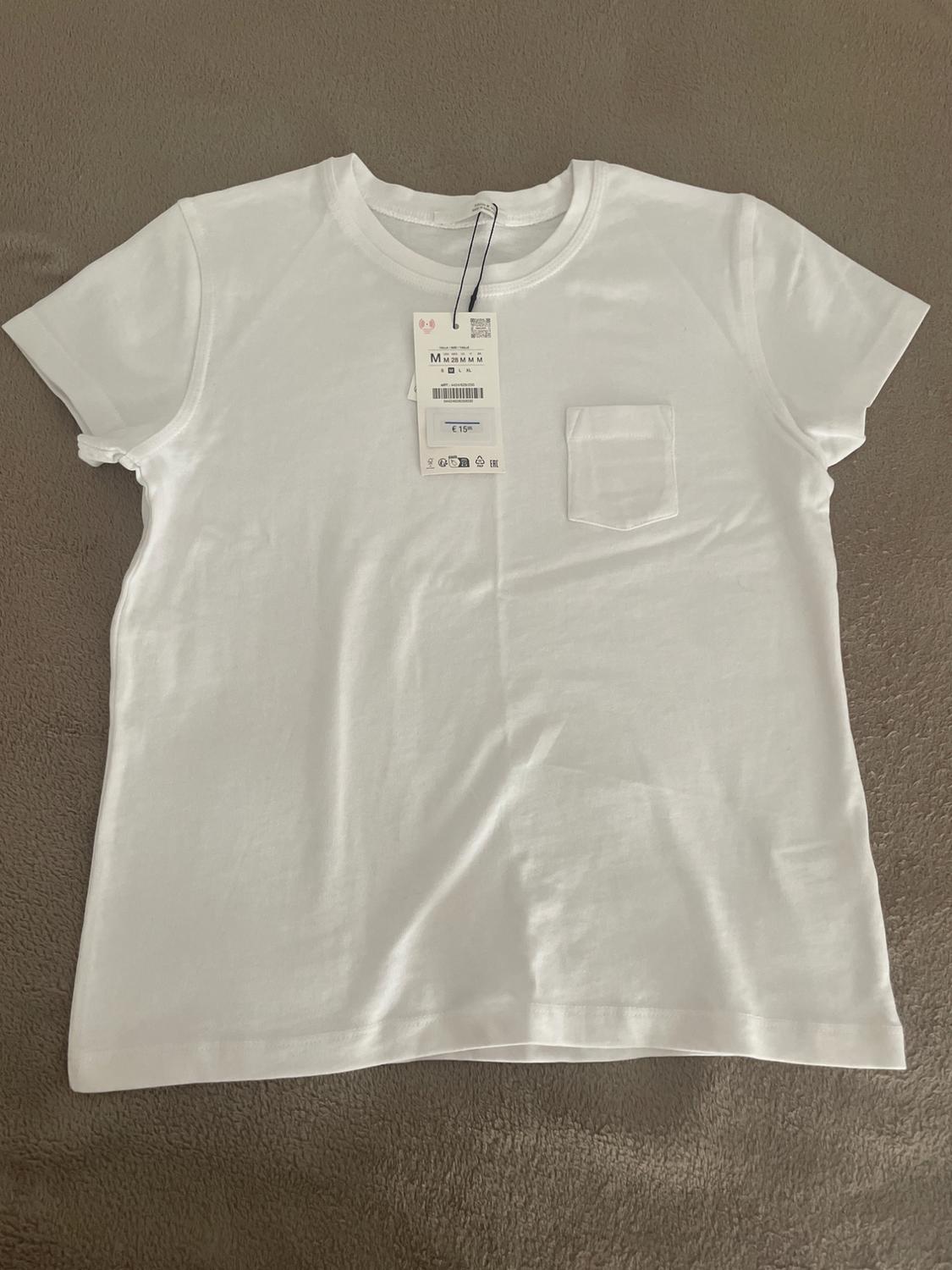 White short-sleeved t-shirt with a front pocket and a label displaying the text 'M' and '€ 15', laid out on a textured beige surface.