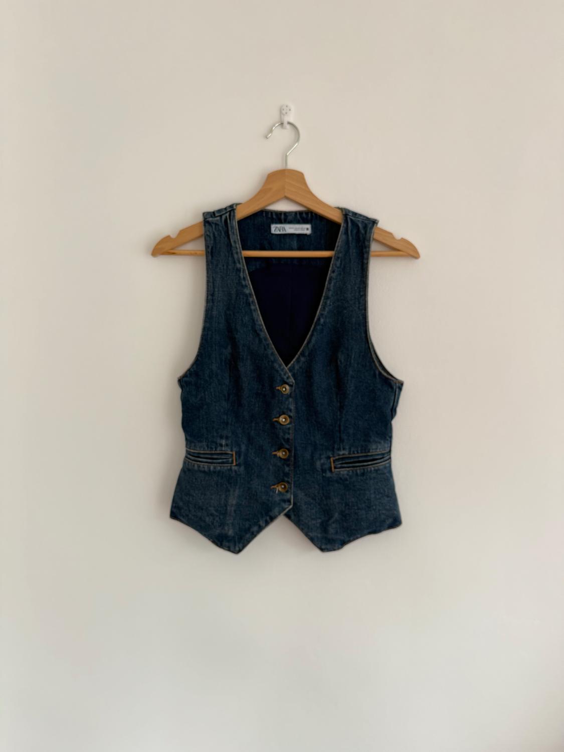 Blue denim waistcoat hanging on a wooden hanger against a white wall. Label visible: "Zara Woman".
