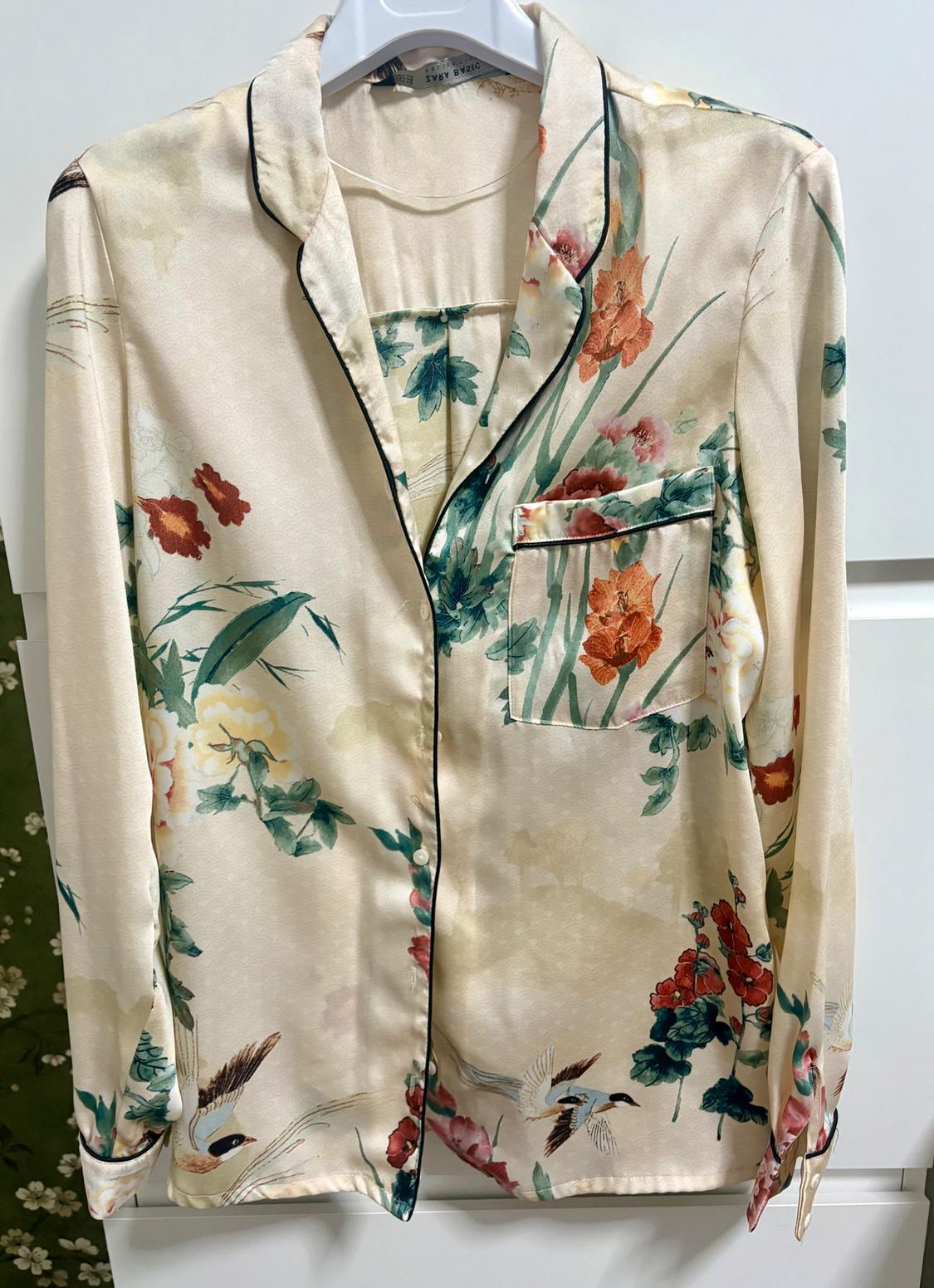 Satin shirt with a floral and bird print in green and orange tones, hanging on a white hanger.