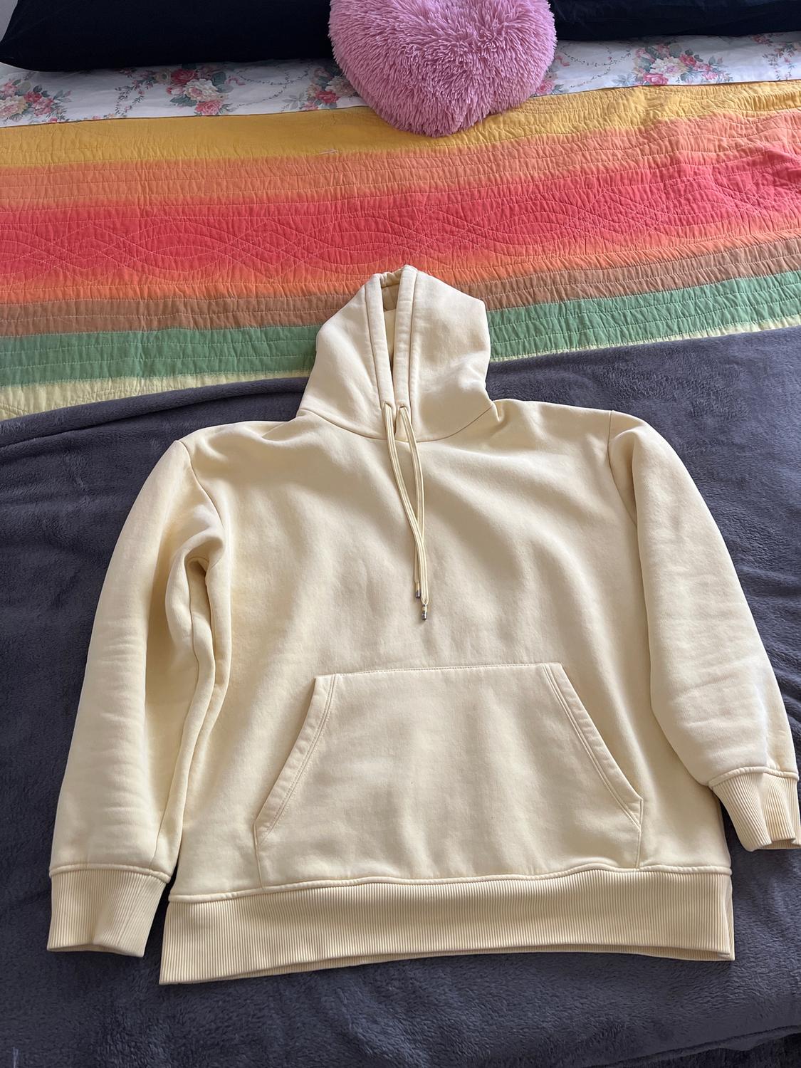 Beige hoodie with a front pocket on a bed with a multicoloured striped bedspread and a pink heart-shaped cushion.