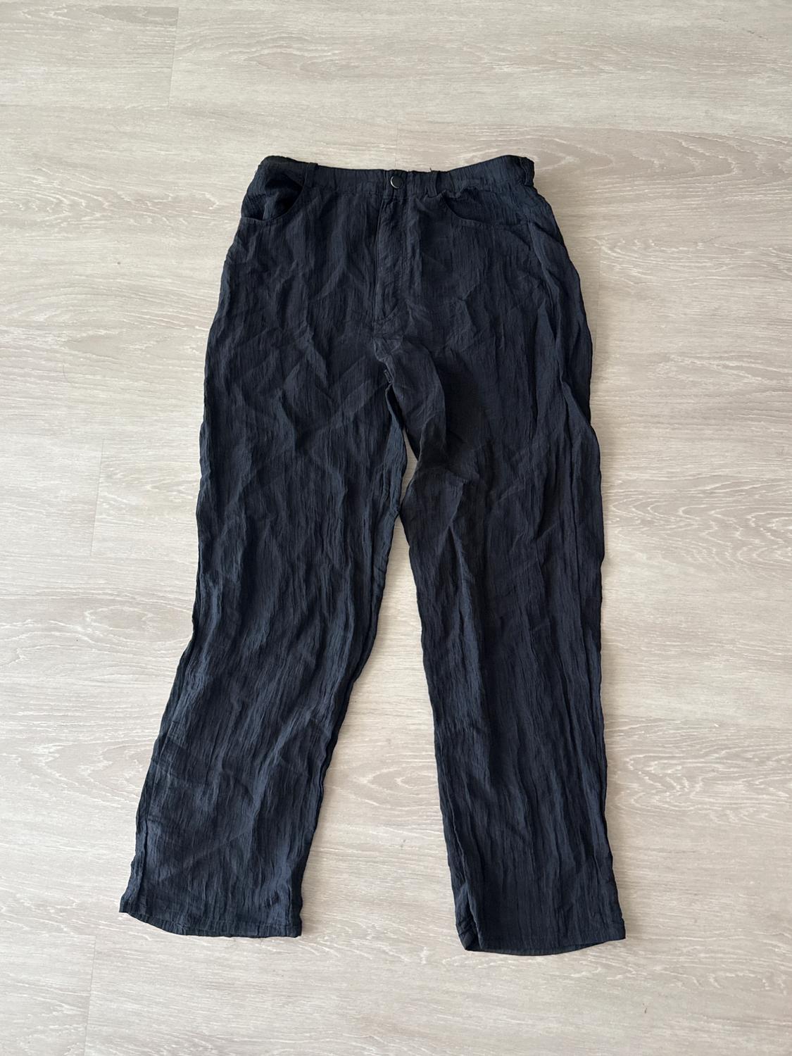 Casual black pants with a crinkled texture and side pockets, laid out on a light wood floor in a relaxed composition.