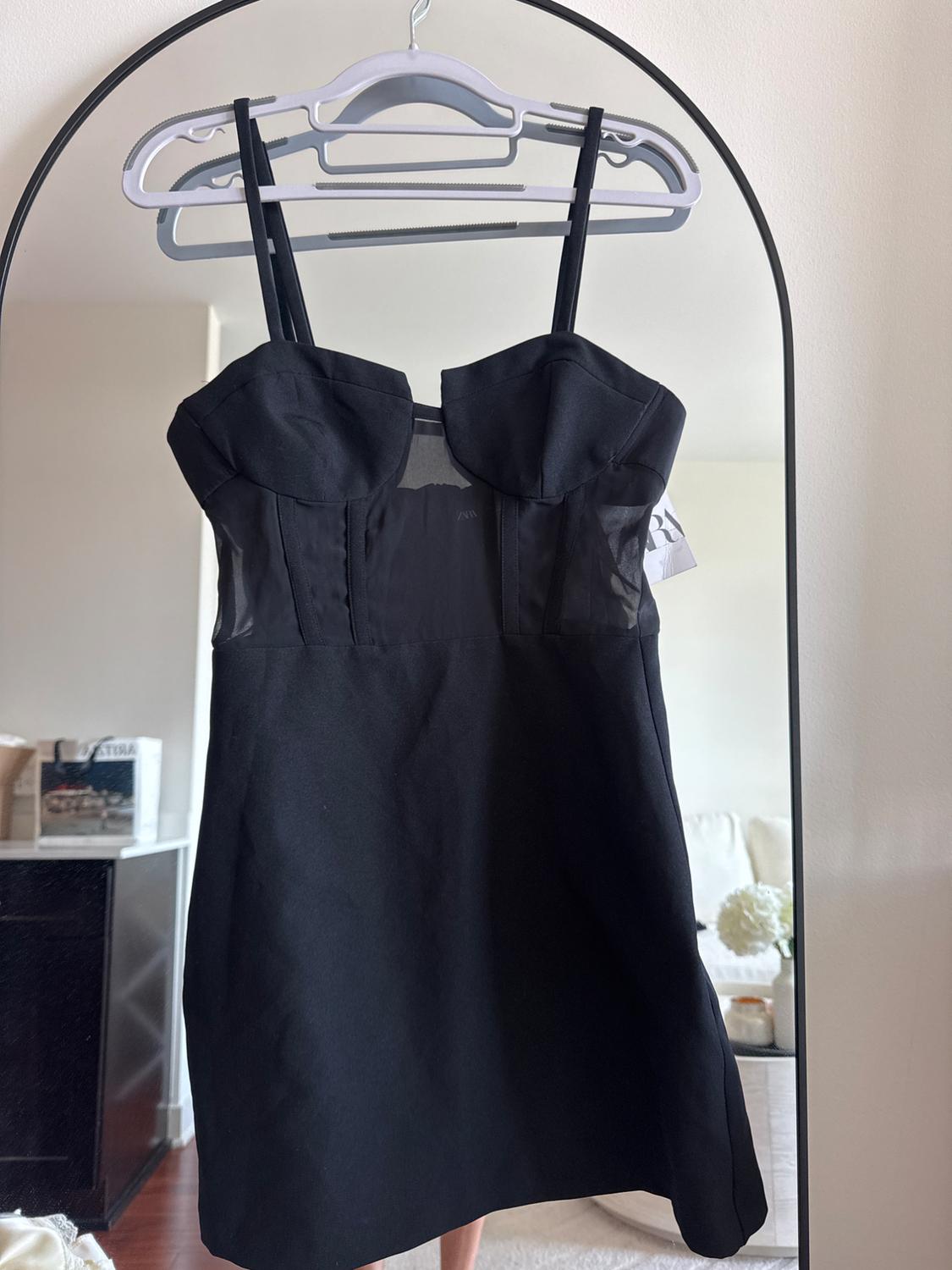 Black dress with spaghetti straps hanging on a white hanger in front of a mirror. Room background with sofa and white flowers.