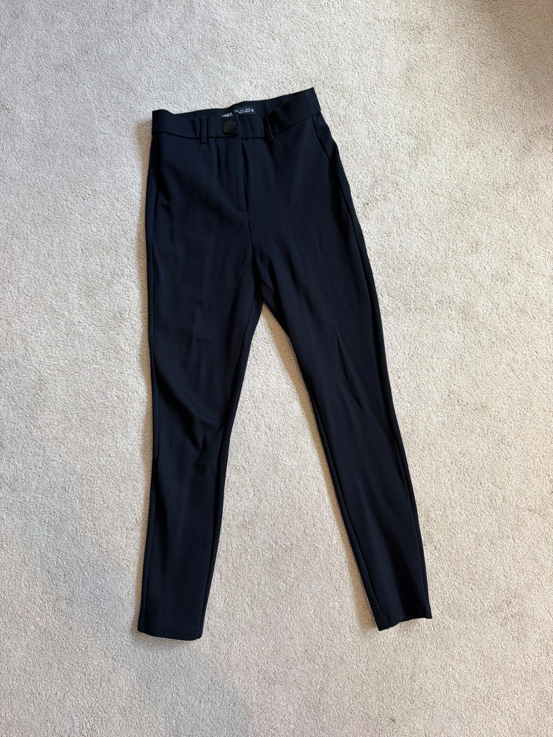 Slim-fit black trousers with a front button, laid out on a light-coloured textured rug. Simple, relaxed-style composition.