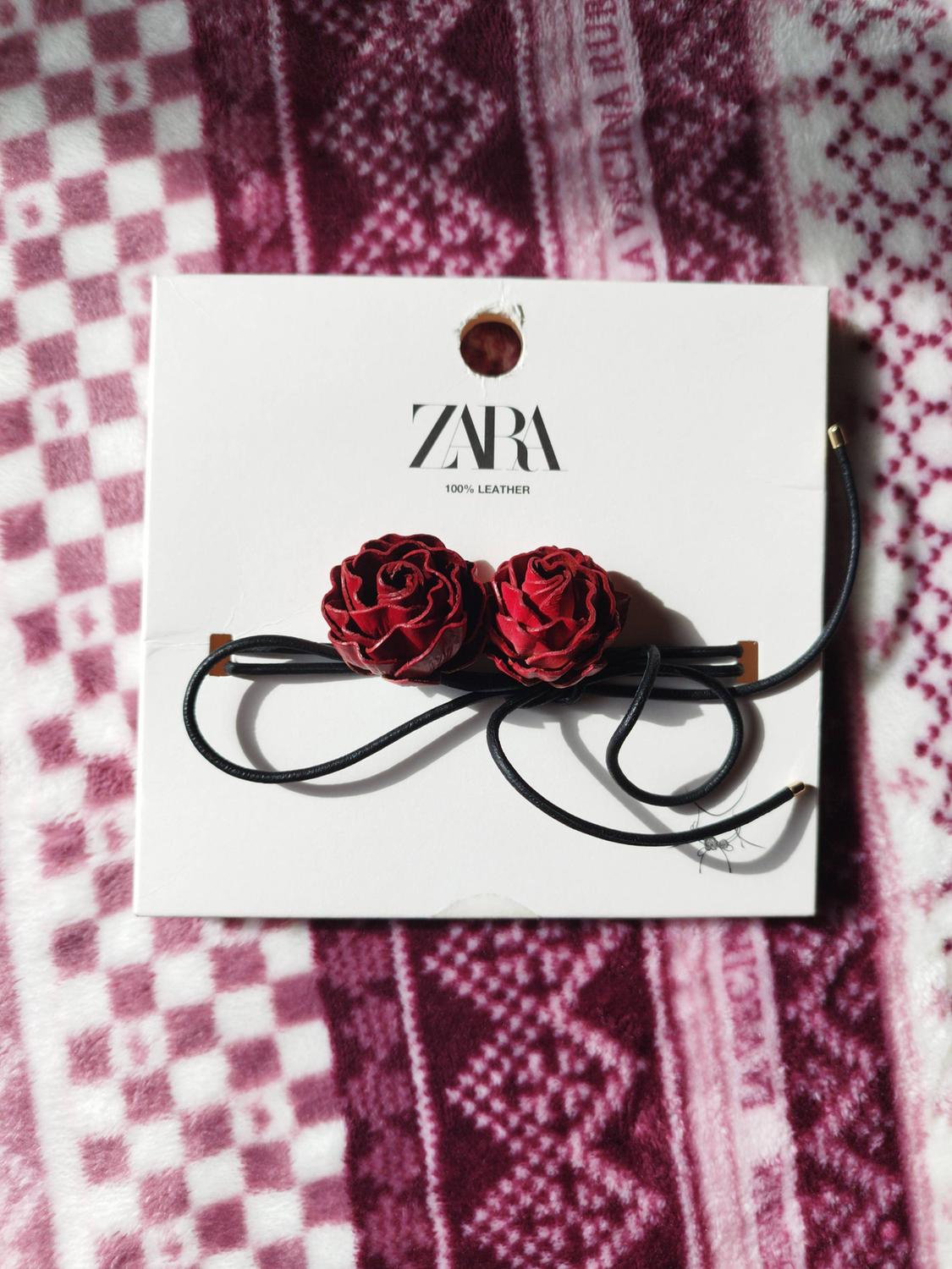 Black cord accessory with two red roses on its tag featuring the text ZARA 100% LEATHER, on a pink-toned plaid blanket.