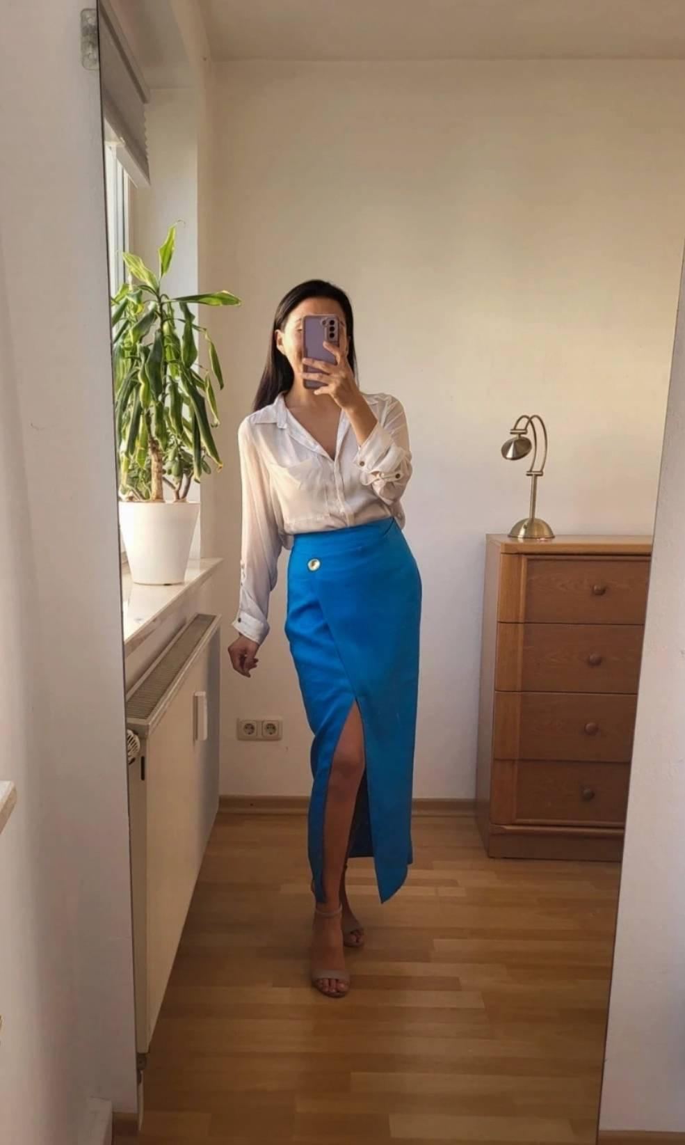Woman in a minimalist room wearing a white blouse and a blue skirt with a slit. She is holding a mobile phone in front of a mirror. Plant and lamp in the background.