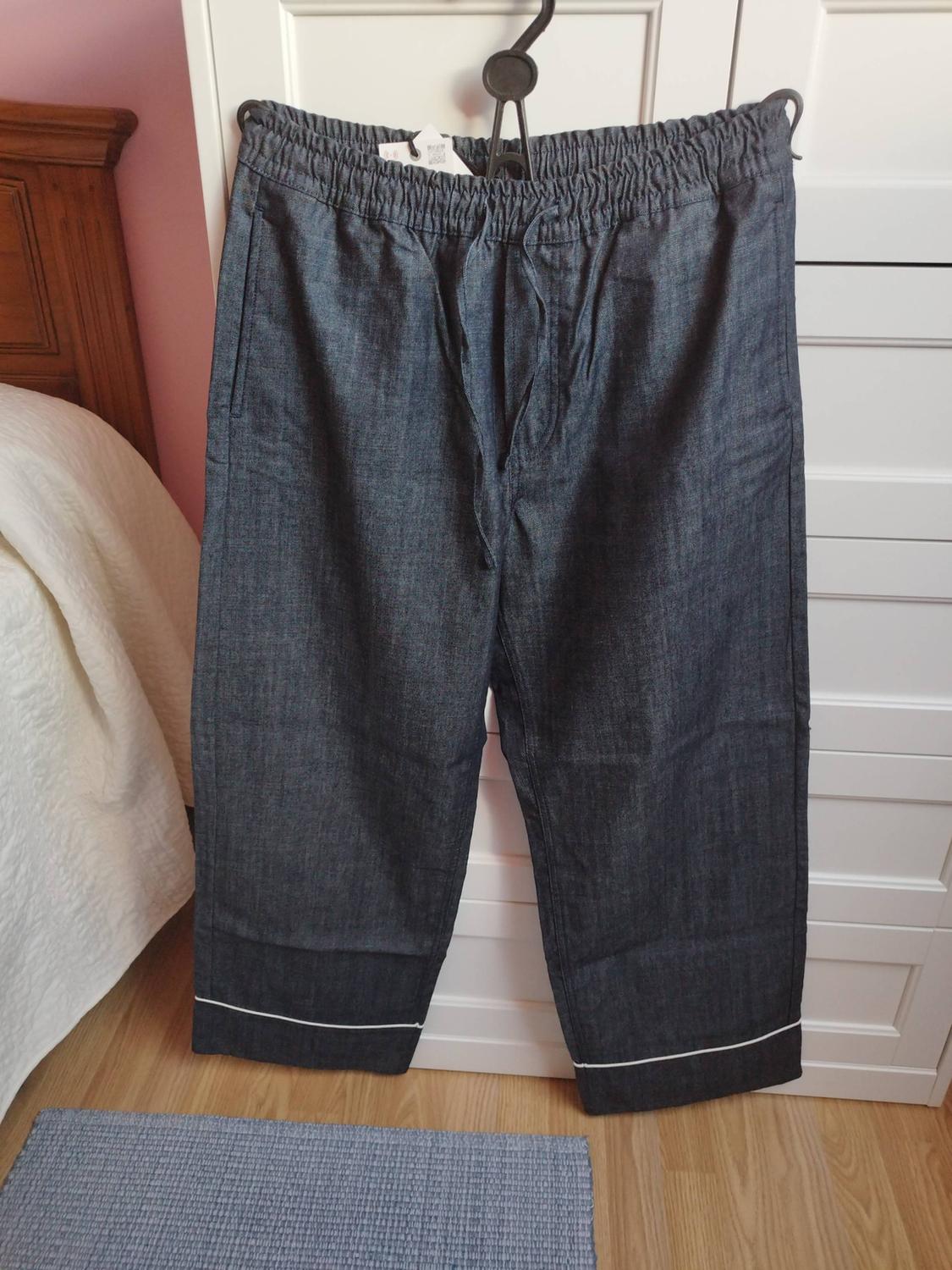 Dark blue trousers with an elasticated waistband, drawstring and white trim, hanging on a hanger in a bedroom with a bed and a white wardrobe.