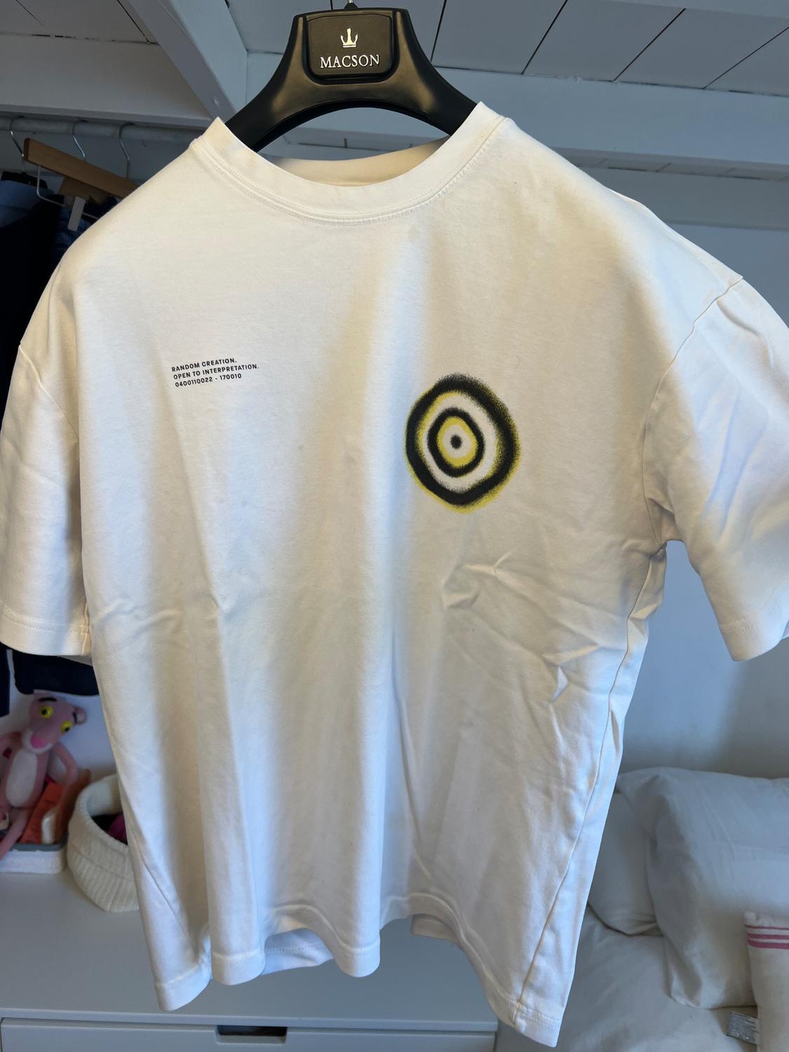 White t-shirt hung on a black hanger with text "RANDOM CREATION. OPEN TO INTERPRETATION." and a black and yellow circular design.