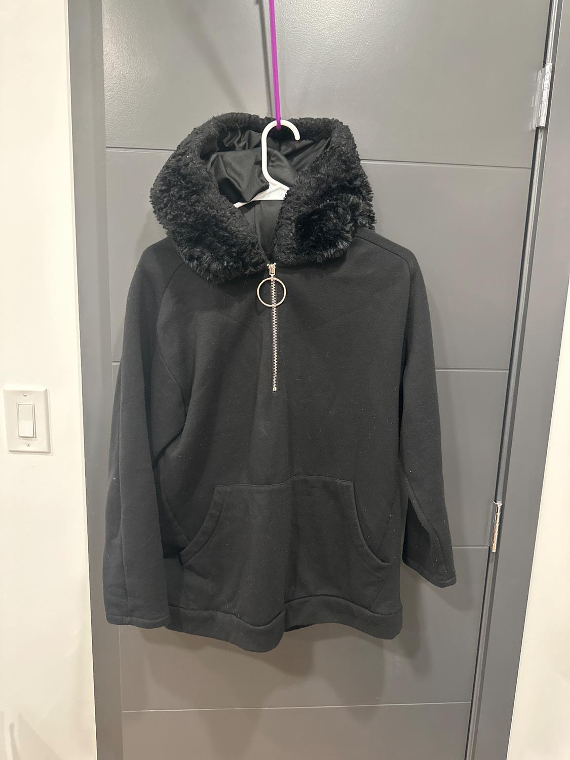Black hoodie with plush hood and circular zipper, hanging on a white hanger over a gray door.