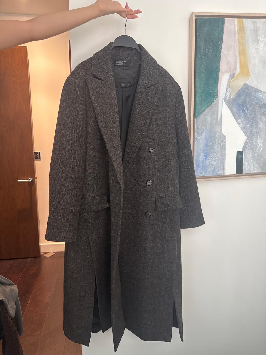 A dark grey coat is hanging on a hanger held by a hand. In the background, there's a wooden door and an abstract painting on a white wall.