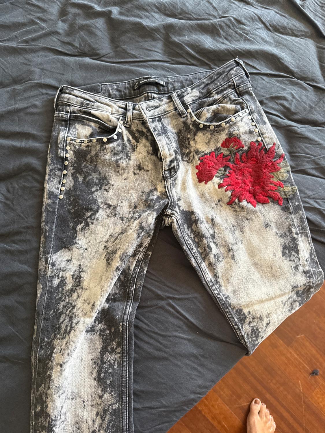 Faded jeans with red floral embroidery on the thigh and studs on the pockets, on a grey bed. Foot visible in the corner.