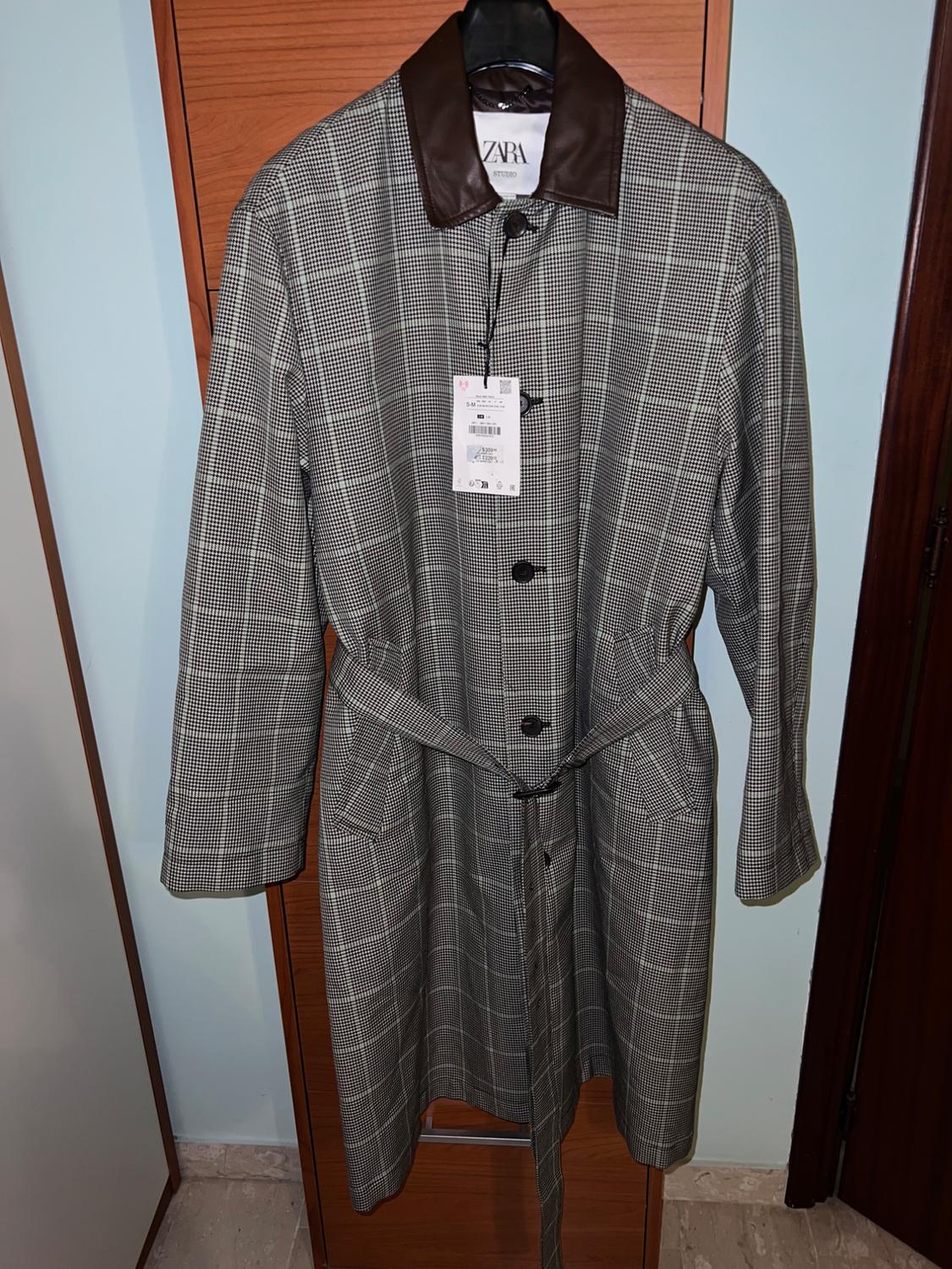 Grey checked coat with a belt and brown collar, hanging on a door. Visible label with the text "ZARA".