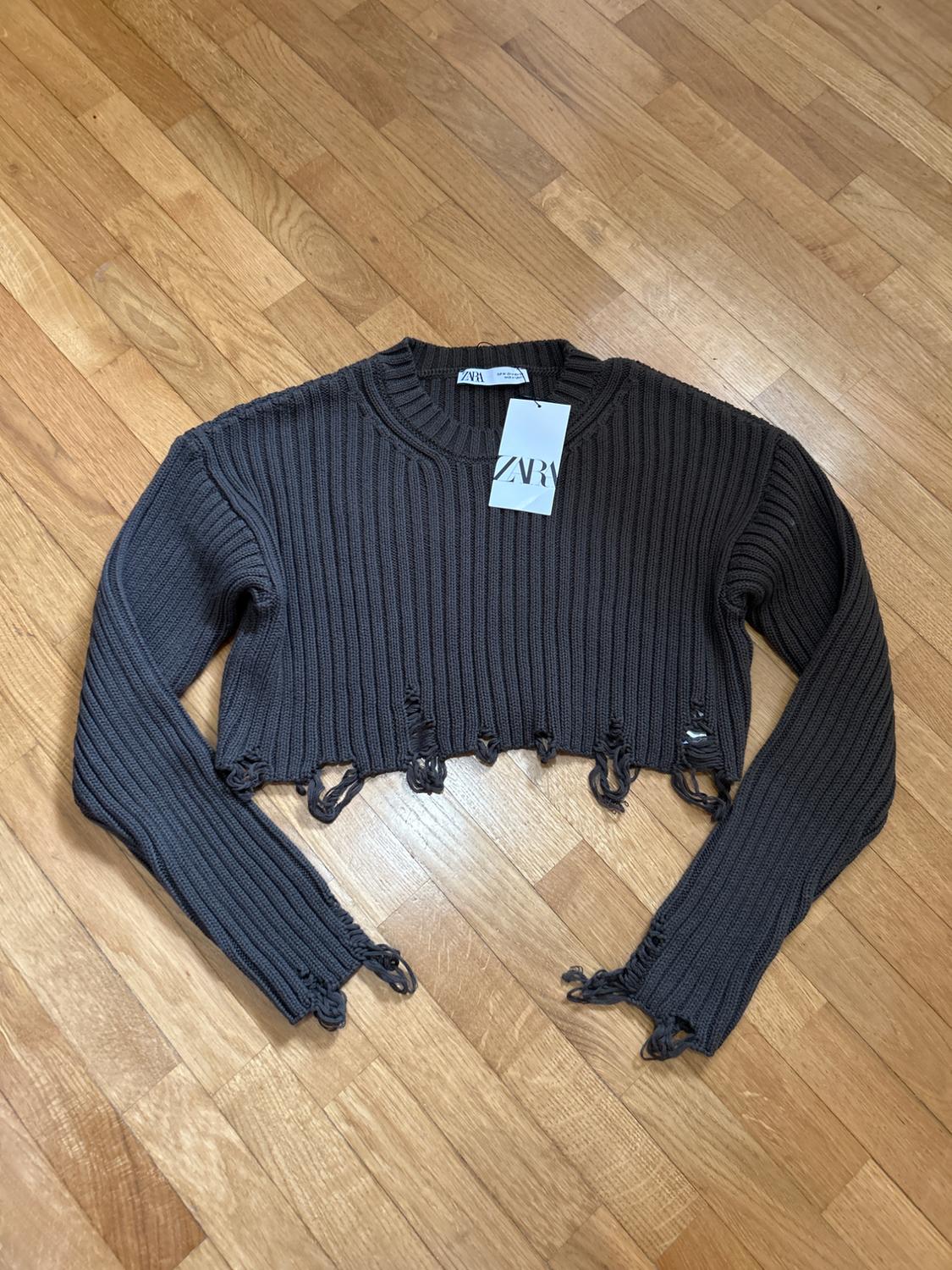 Dark grey ribbed knit cropped jumper, long sleeves, frayed hem and cuffs, on a wooden floor. Labels with ZARA text.