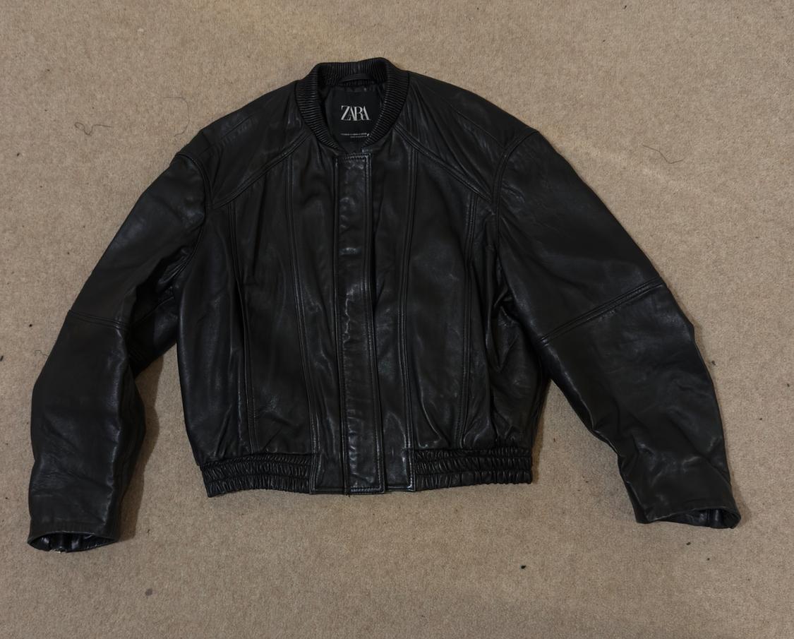 Black leather jacket with elasticated waist and cuffs, on a beige background. Visible label with the text "ZARA".