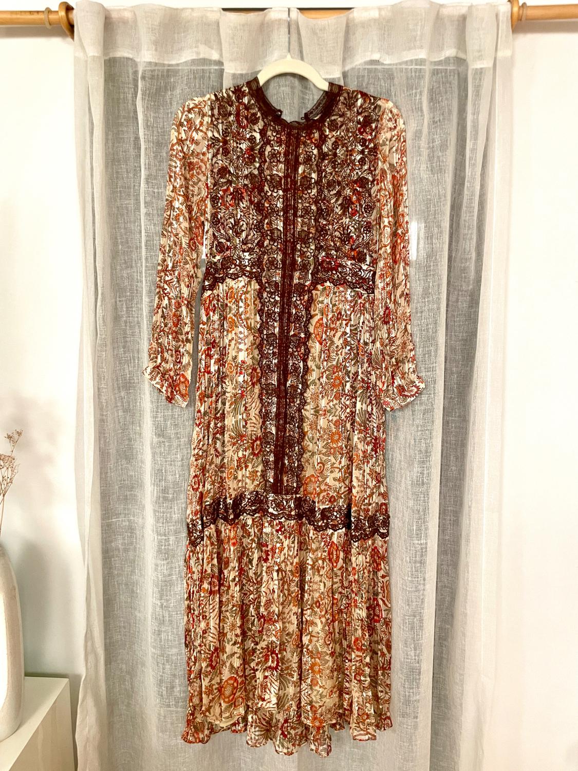 Long floral print dress in brown and red tones, hanging on a hanger in front of a translucent white curtain.