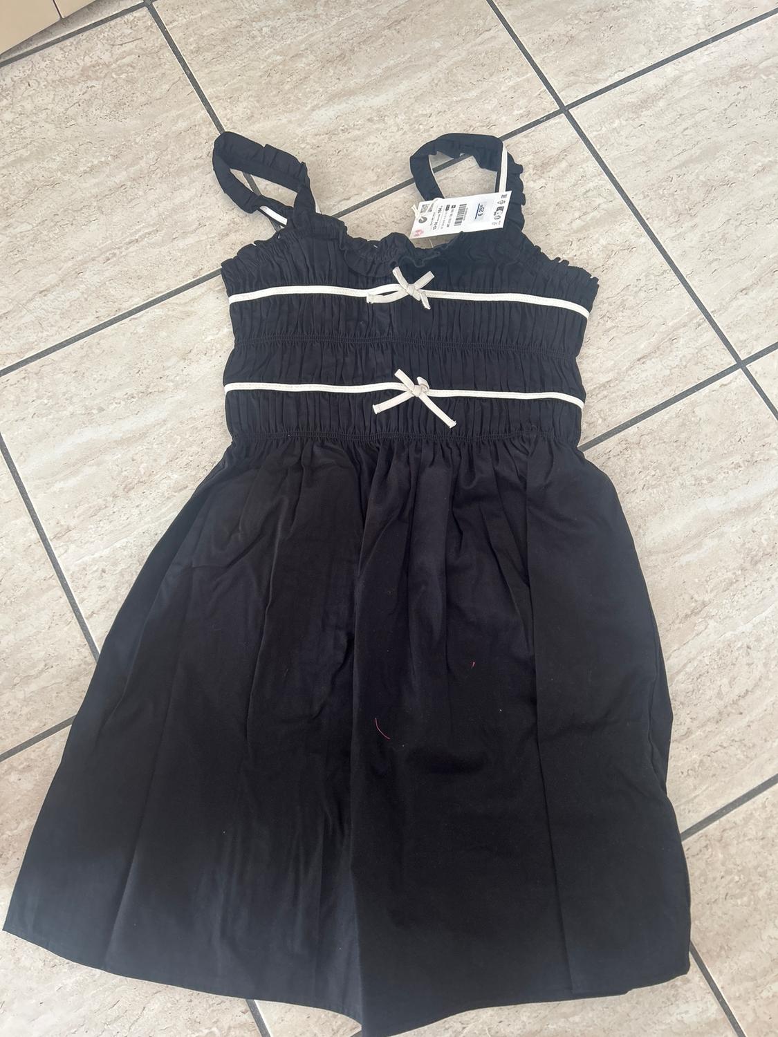 Black dress with straps and white bow details, placed on a light tiled floor. Label visible on the strap.