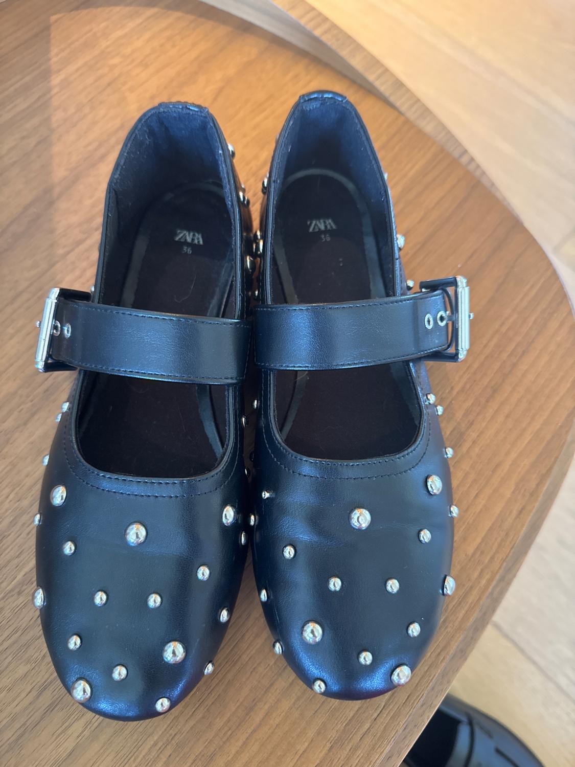 Black shoes with sparkly studs and silver buckles on a wooden table. Visible text: "ZARA 36".