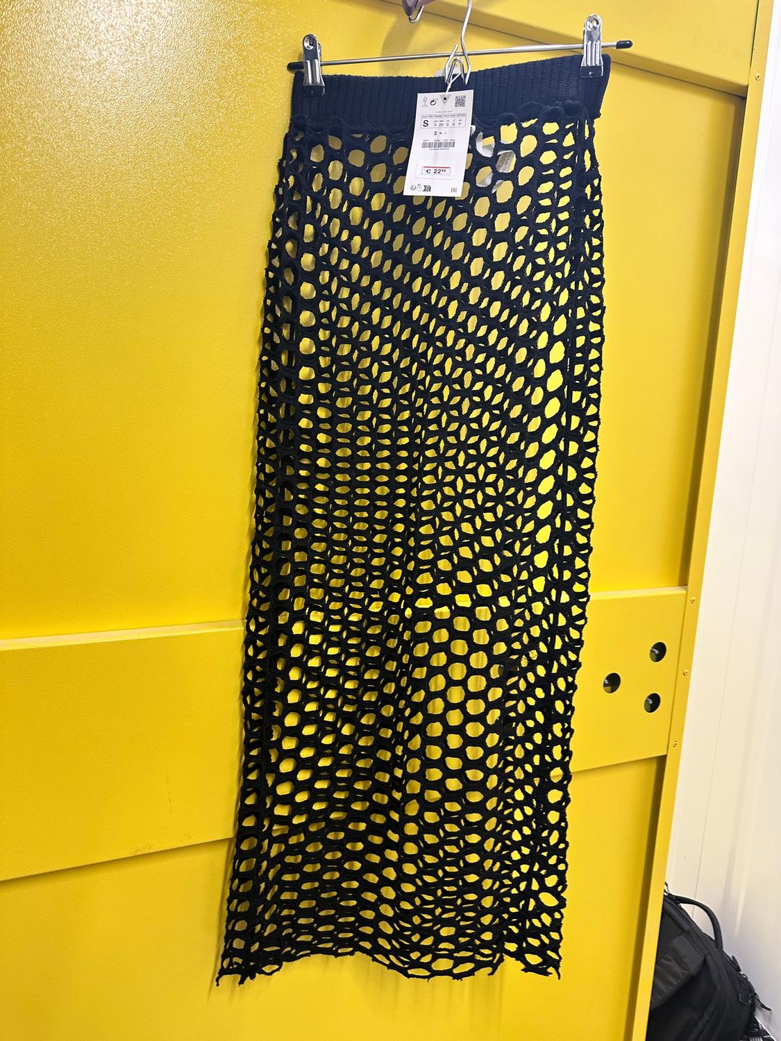 Black mesh skirt hanging on a hanger against a bright yellow background. Visible tag with product details.
