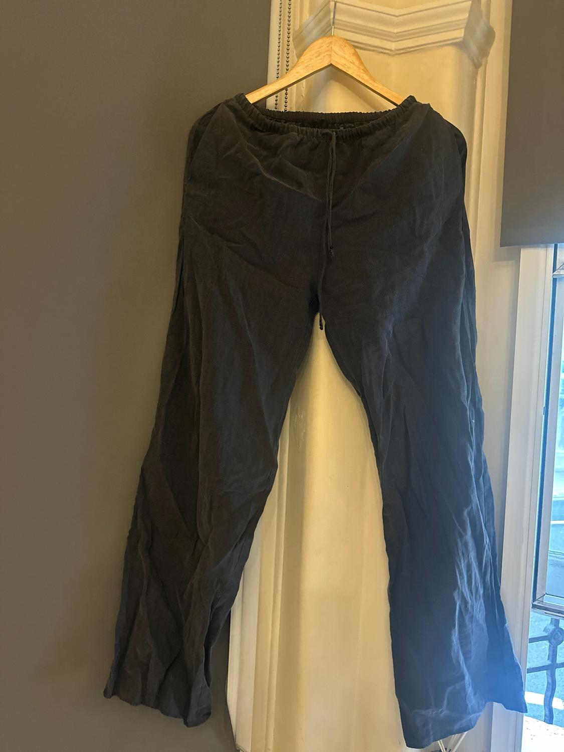 Dark, loose-fitting trousers hanging on a wooden hanger against a grey and white wall beside a window. Casual and relaxed atmosphere.