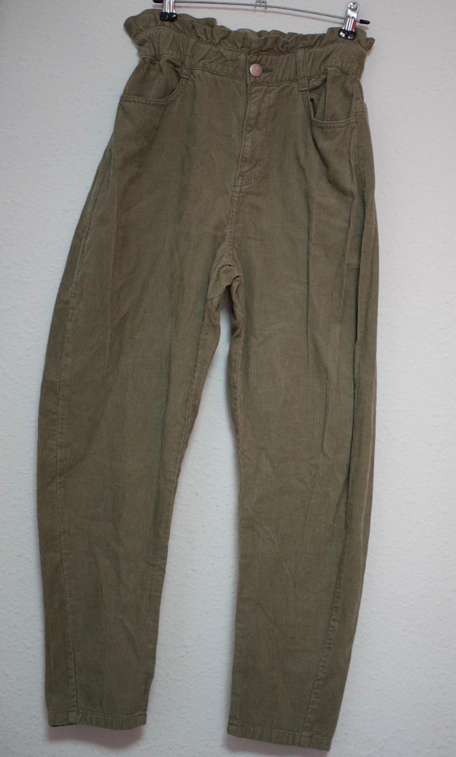 Olive green corduroy trousers hanging on a hanger against a white background. High-waisted, elasticated design with frills, a central button and pockets.