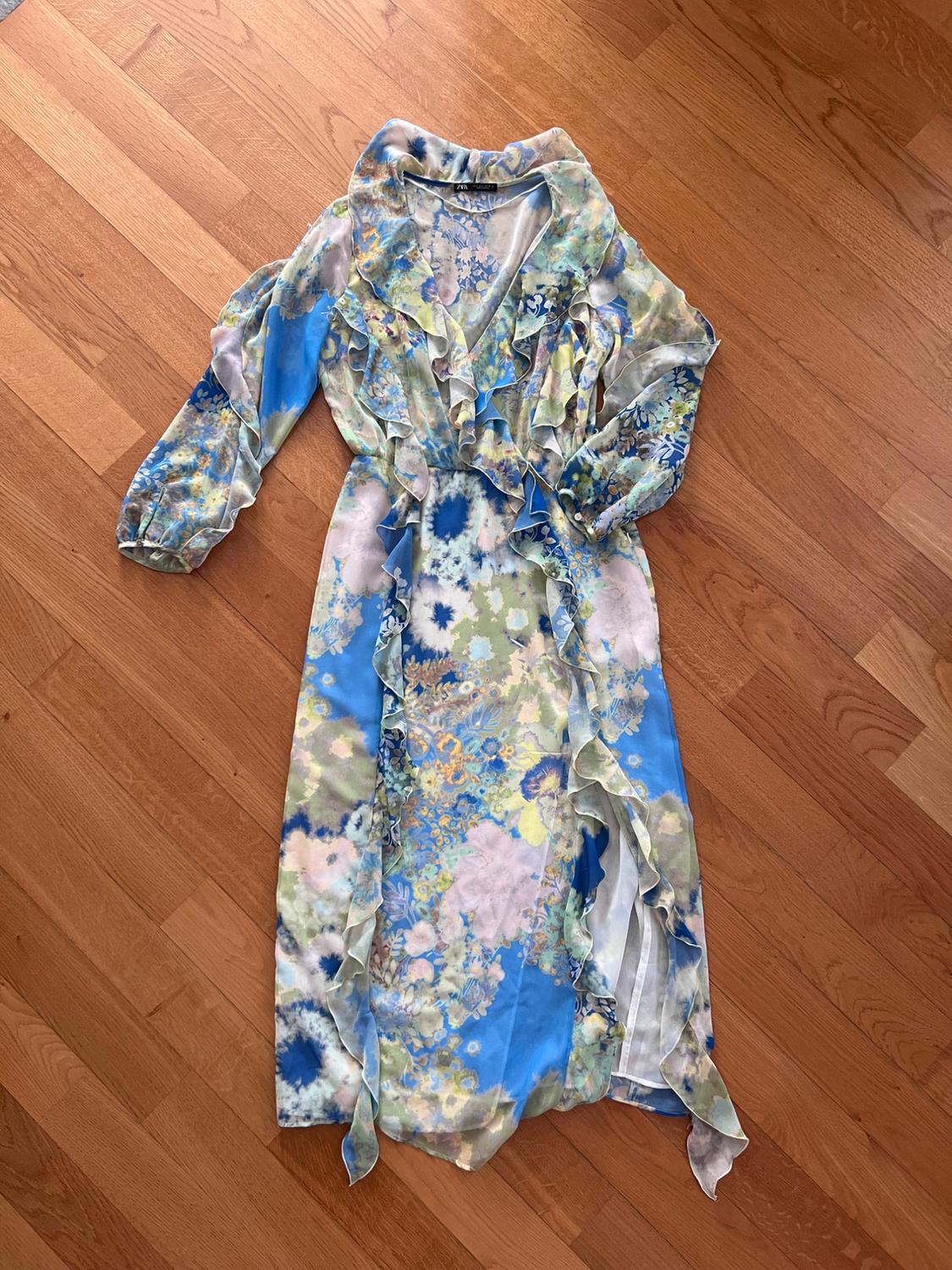 Long-sleeved maxi dress with a floral print in blue and green tones, V-neck, and frills. Laid out on a wooden floor.