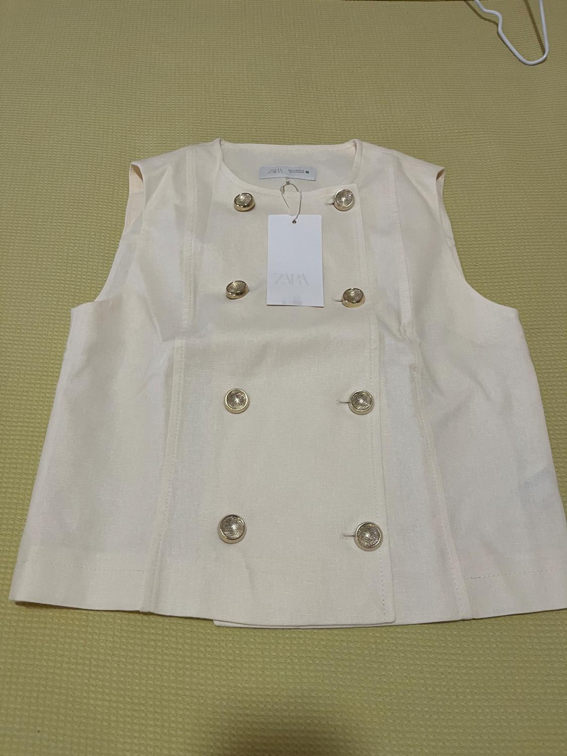 Cream sleeveless waistcoat with gold buttons and Zara label, on a beige surface.