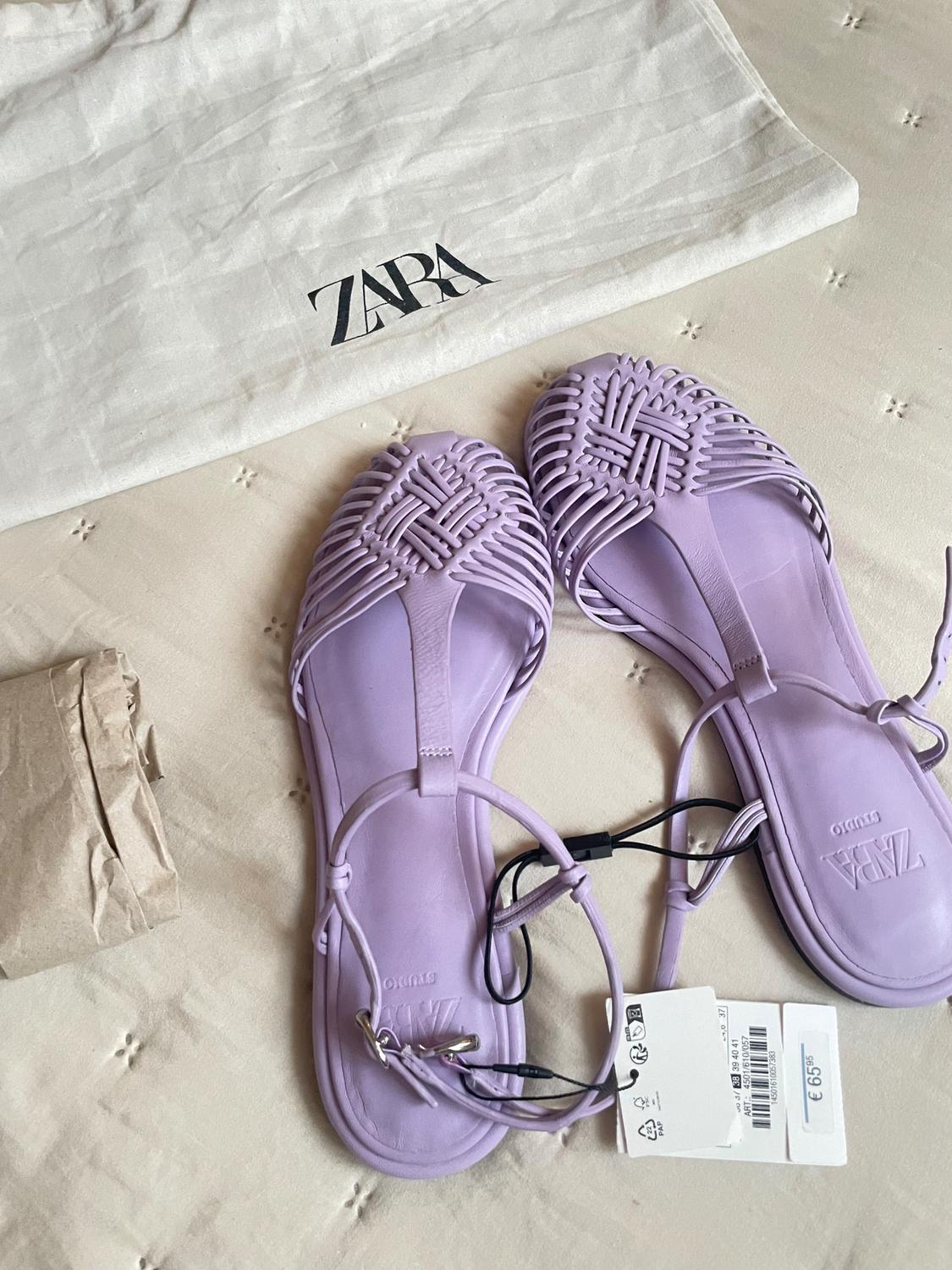 Lilac strappy sandals on white fabric with the "ZARA" logo. Price tag of €69.95. Brown paper next to it.