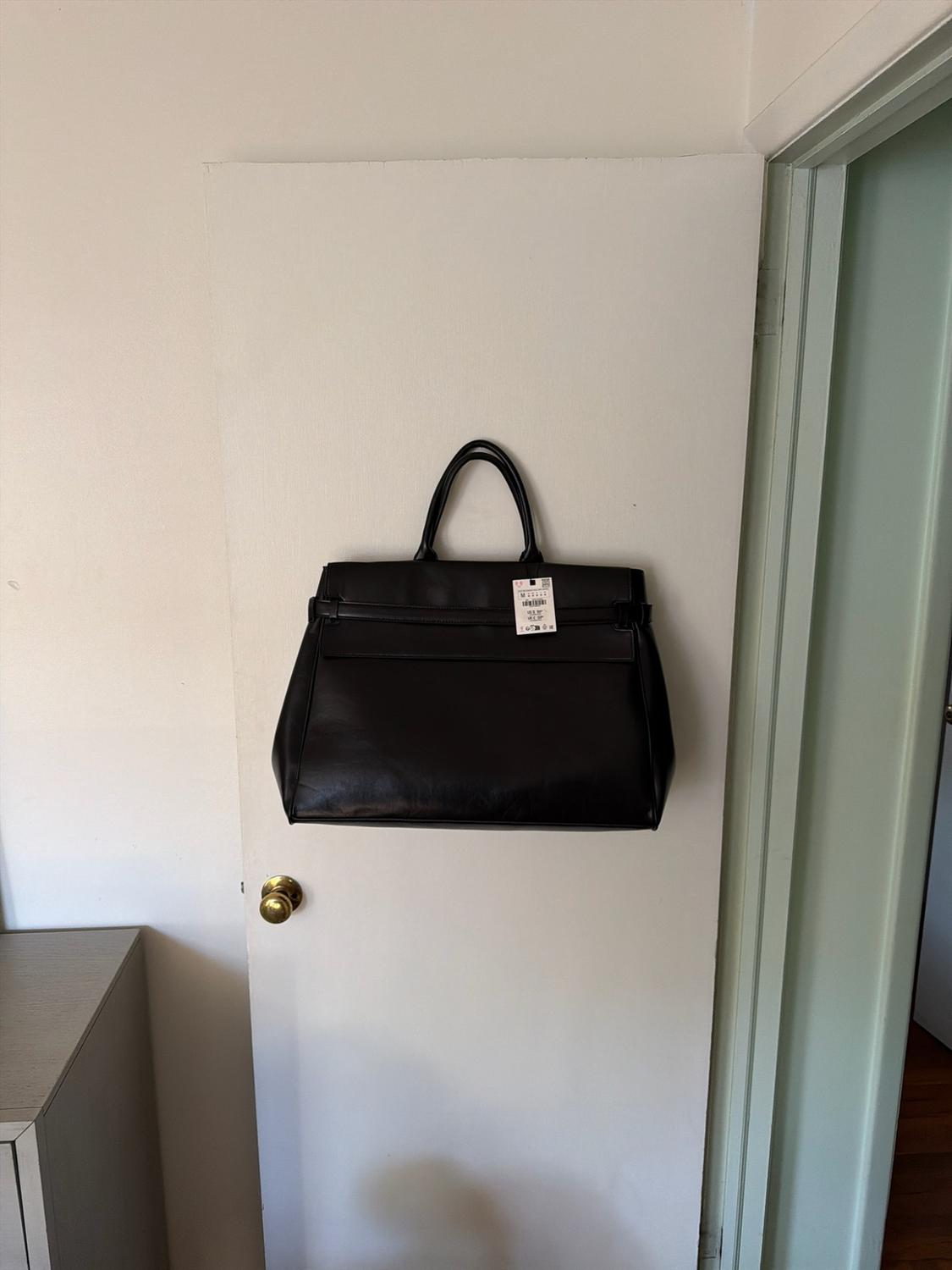 Large black bag with handles and a tag hanging on a white door with a golden knob in a simple interior.