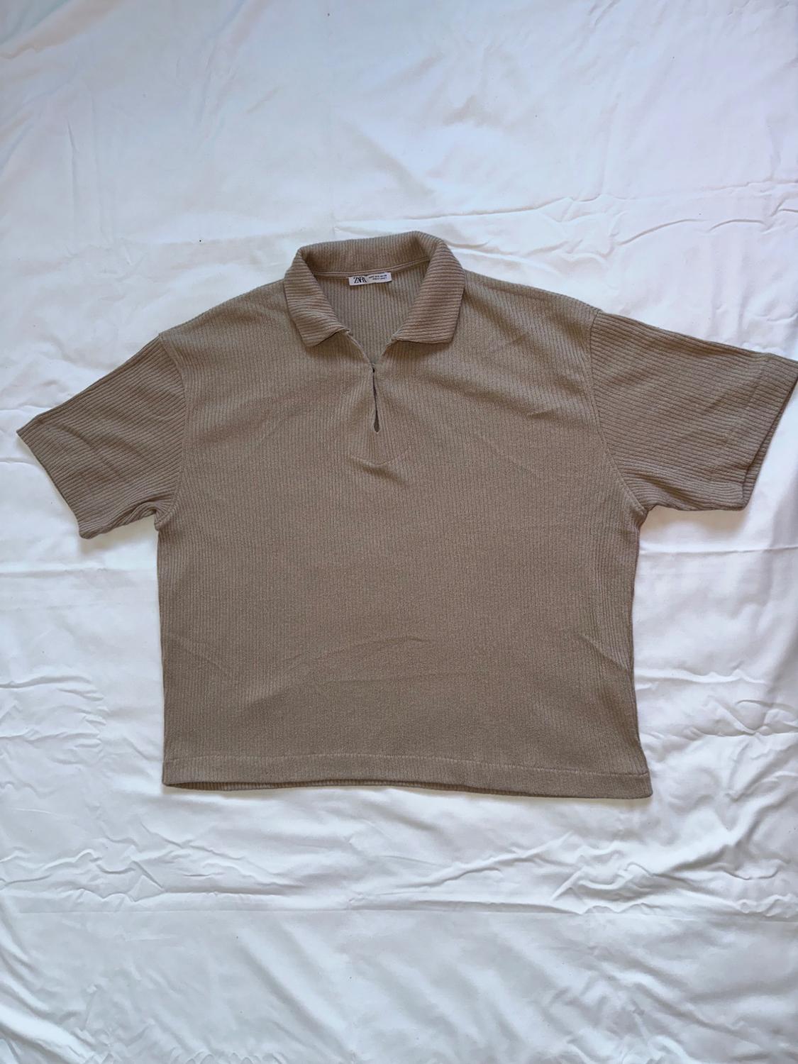Beige short-sleeved polo shirt with ribbed texture, placed on a wrinkled white surface. Label visible at the neck.
