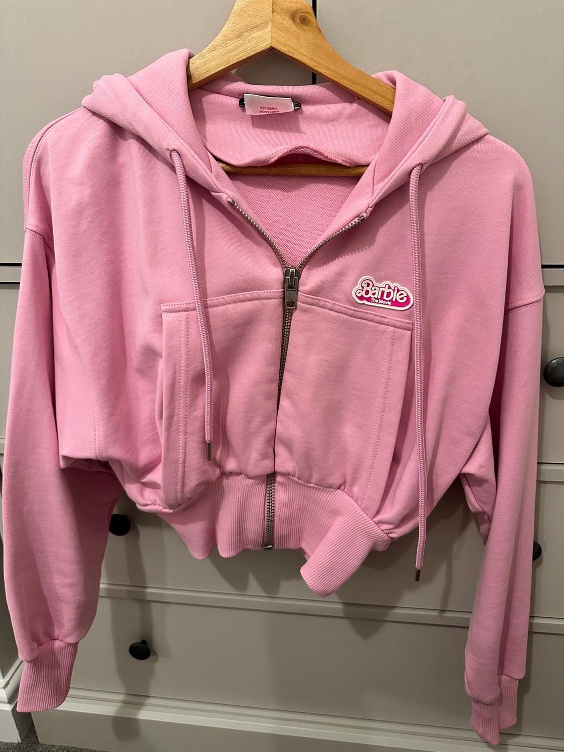 Pink hoodie with a zip, hanging on a wooden hanger. "Barbie The Movie" logo on the chest. Chest of drawers in the background.