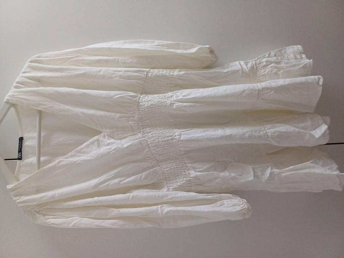 White, long-sleeved dress with a gathered waist, hanging on a white hanger against a neutral background. Label visible.