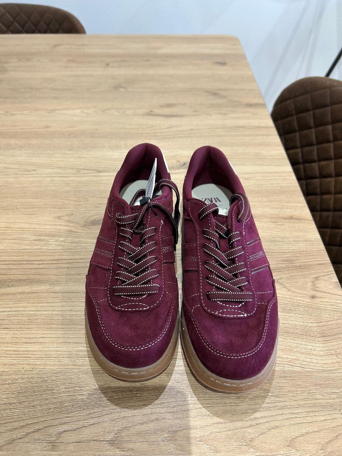 Burgundy lace-up trainers on a light wooden table. Background with brown upholstered chairs. Label visible inside.