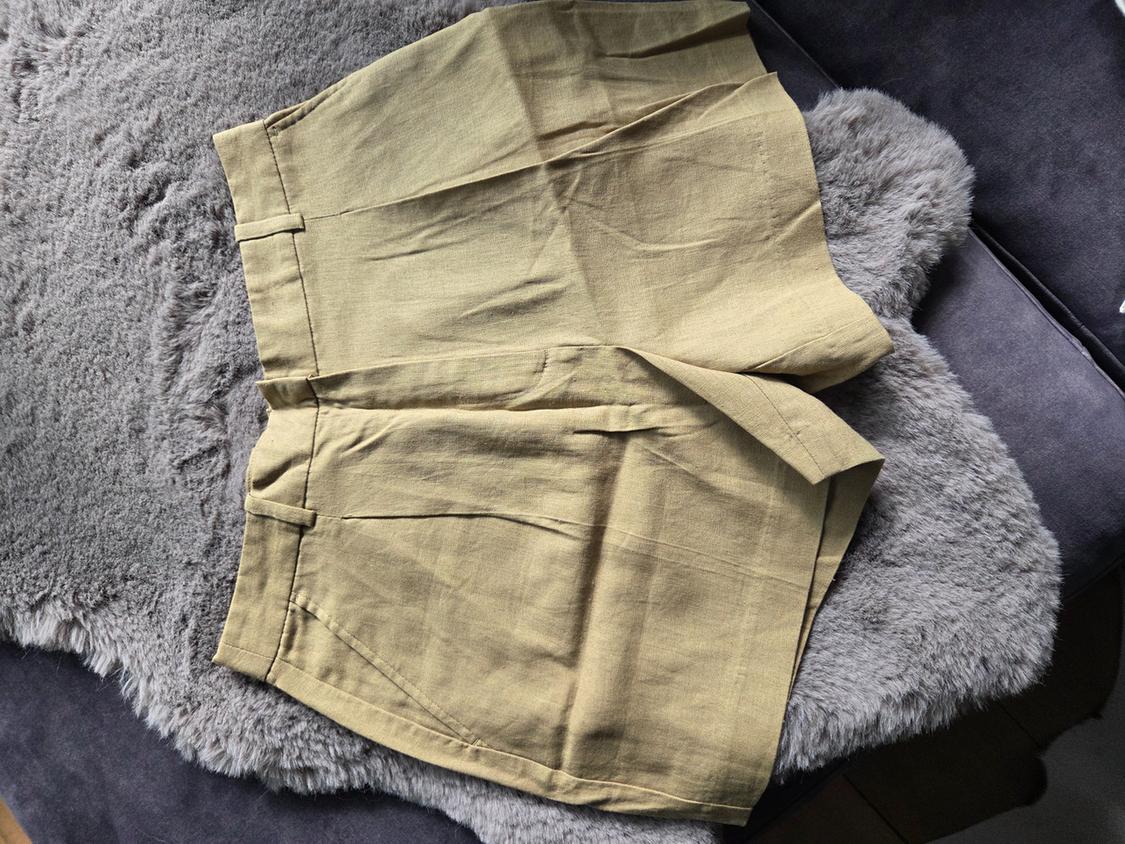 Khaki shorts with pleats and pockets, displayed on a grey fur blanket in a relaxed setting.