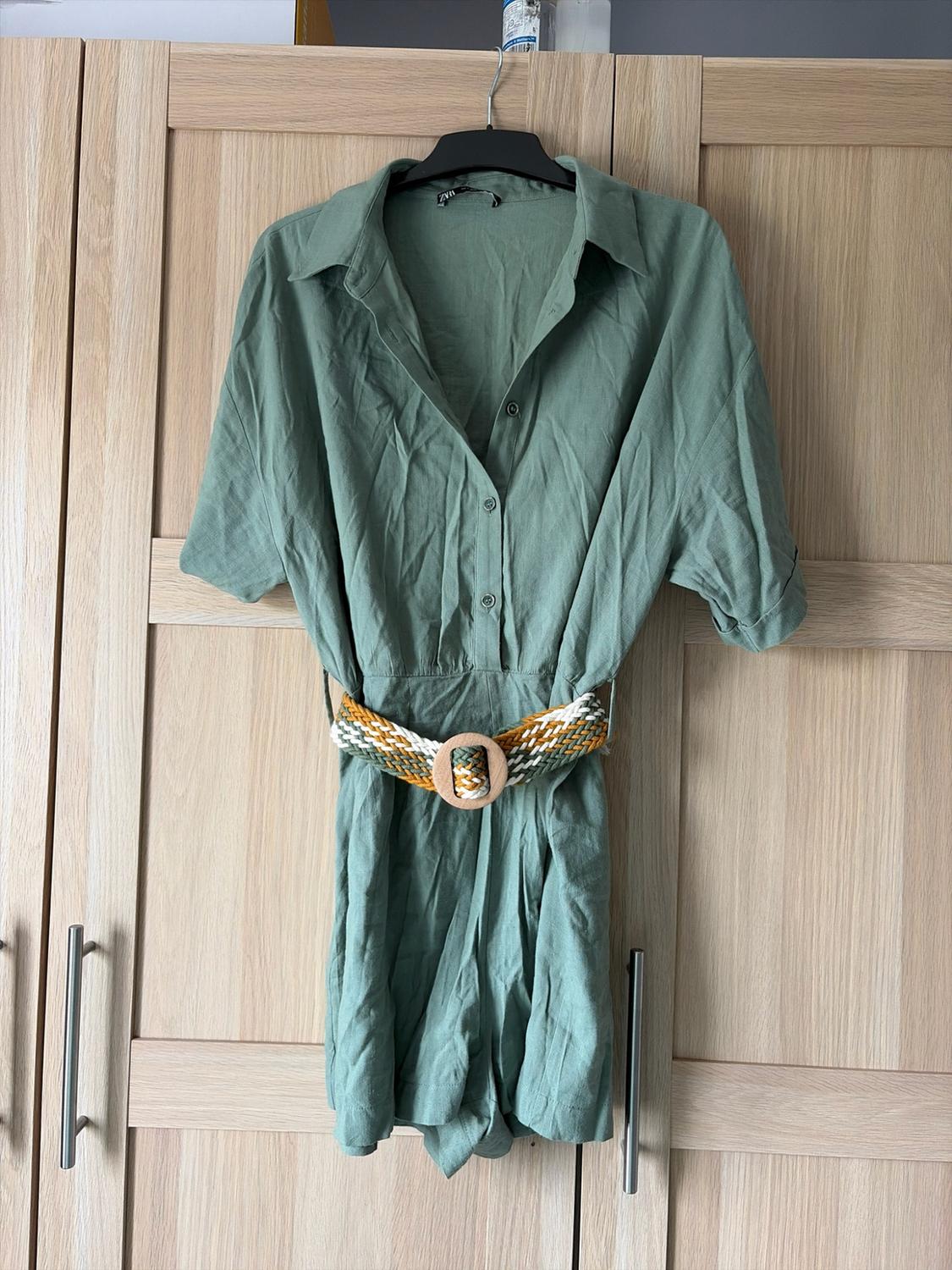 Light green casual playsuit with buttons and a braided belt. Hanging in a light wood wardrobe. Text: ZARA.