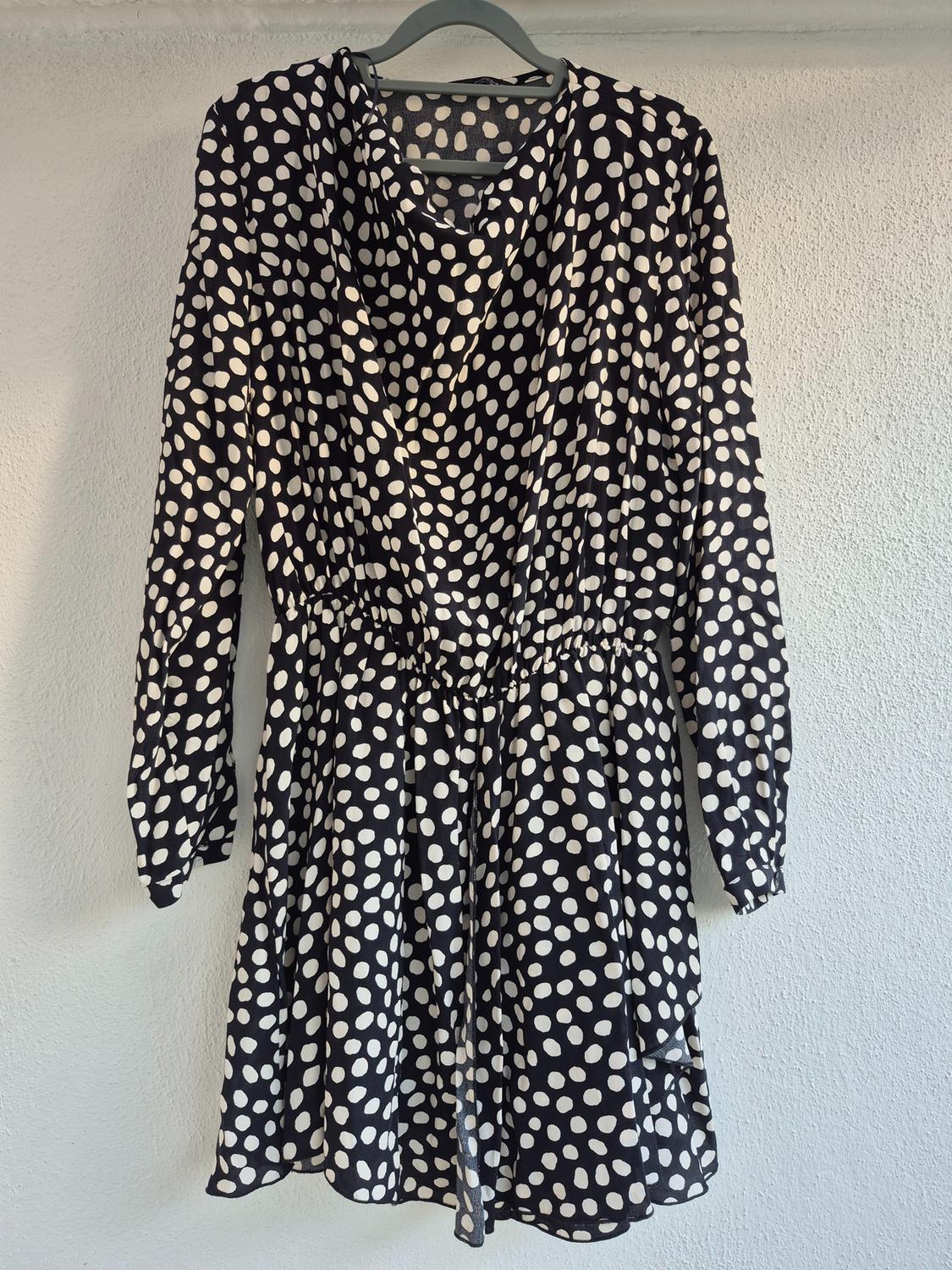 Black dress with white polka dots hanging on a grey hanger, with long sleeves and a fitted waist, against a white background.