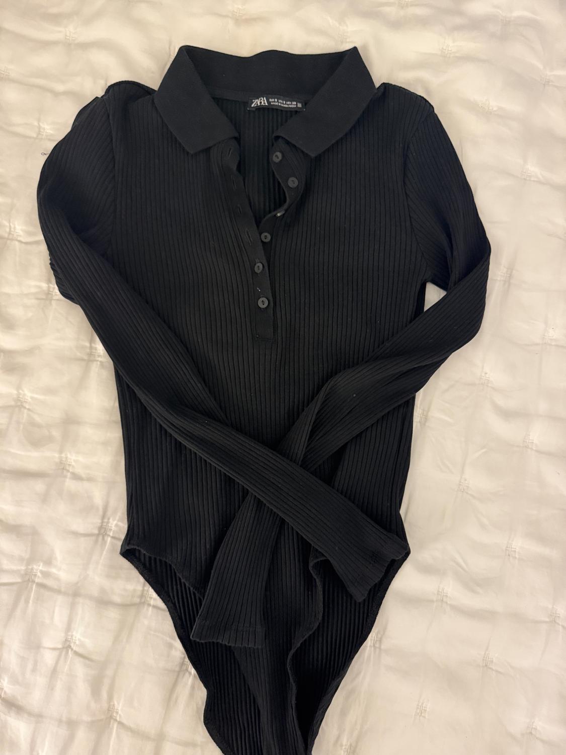 Black long-sleeved bodysuit with a polo neck and buttons, on a white bedspread. Label visible on the neck.