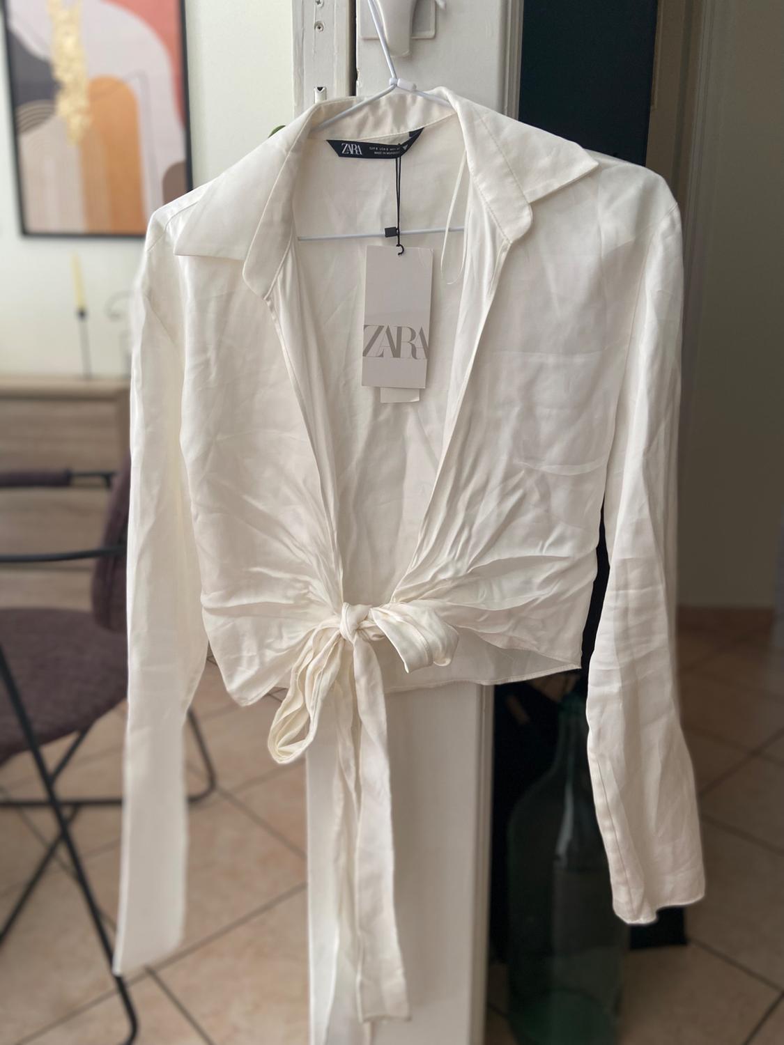 White long-sleeved shirt with front knot hanging on a hanger. Visible label with the text "ZARA". Background with chair and picture.
