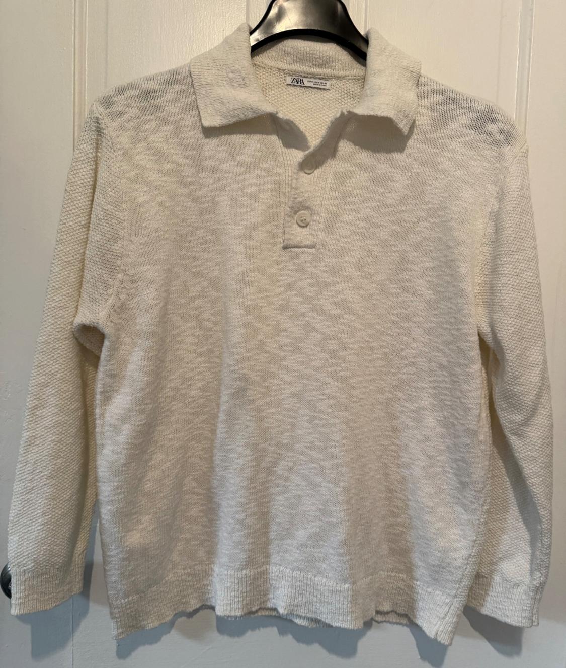 White long-sleeved polo-neck sweater with two buttons, hanging on a black hanger in front of a white door.