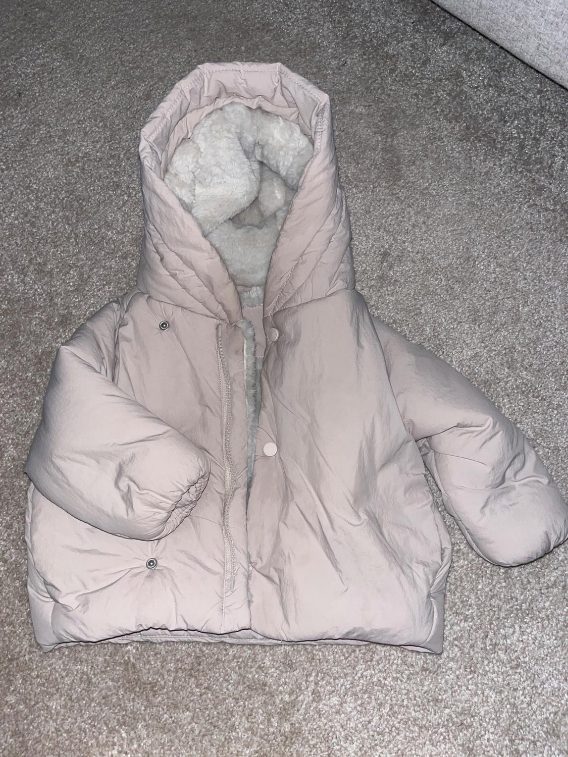 Beige quilted jacket with hood and sheepskin lining, placed on a grey rug.