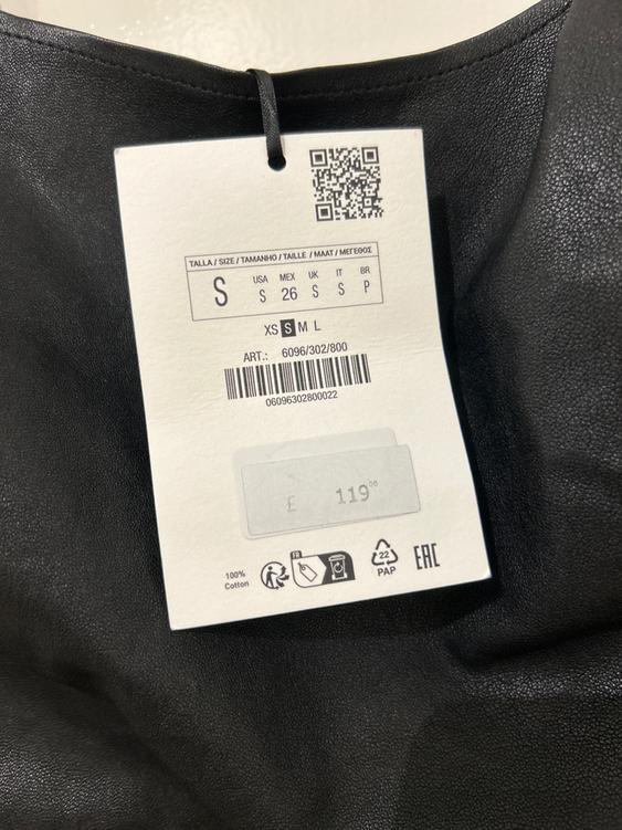 Clothing label on black fabric, shows sizes S, XS, M, L and price £119. Barcode and QR visible. 100% cotton.