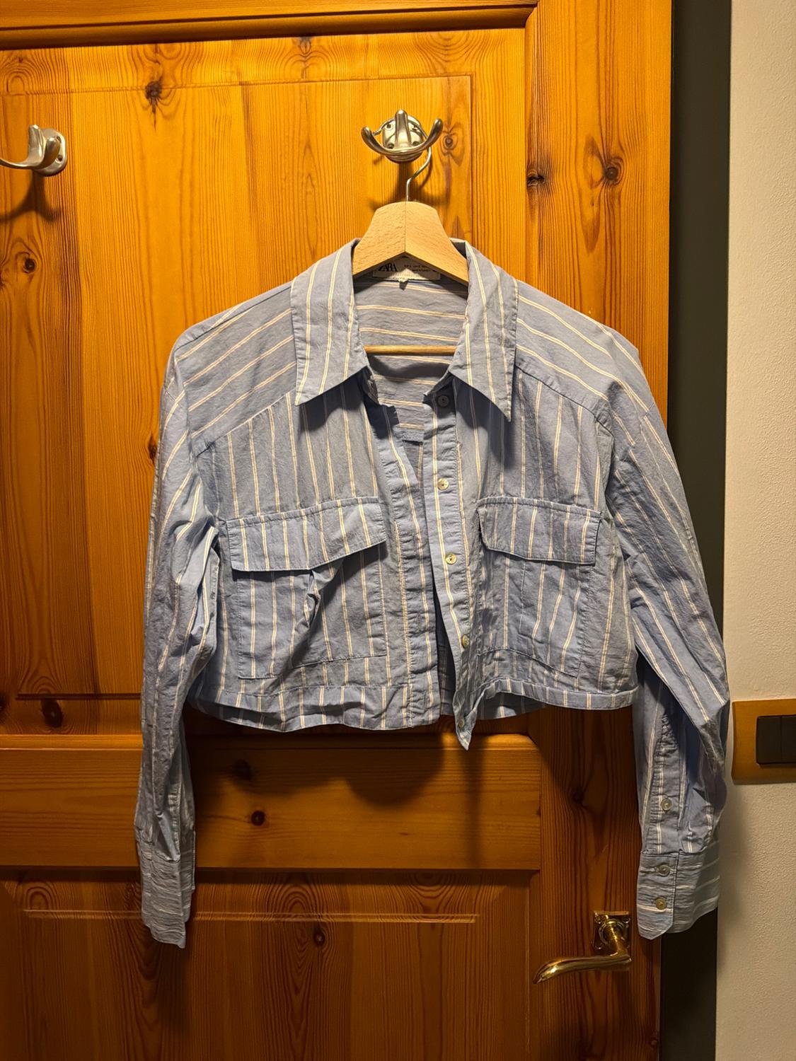 Short-sleeved ZARA shirt with blue and white stripes and pockets, hanging on a coat hanger over a wooden door.