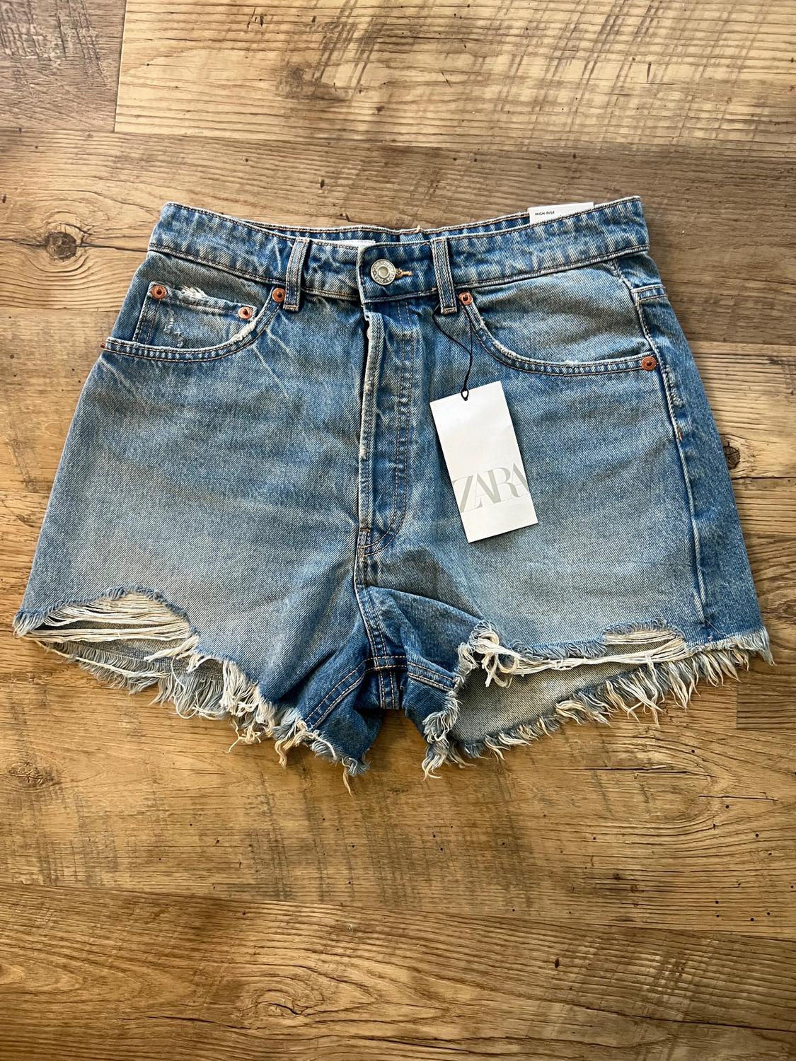 Distressed denim shorts with a Zara tag on a wooden floor. Casual design with frayed edges.
